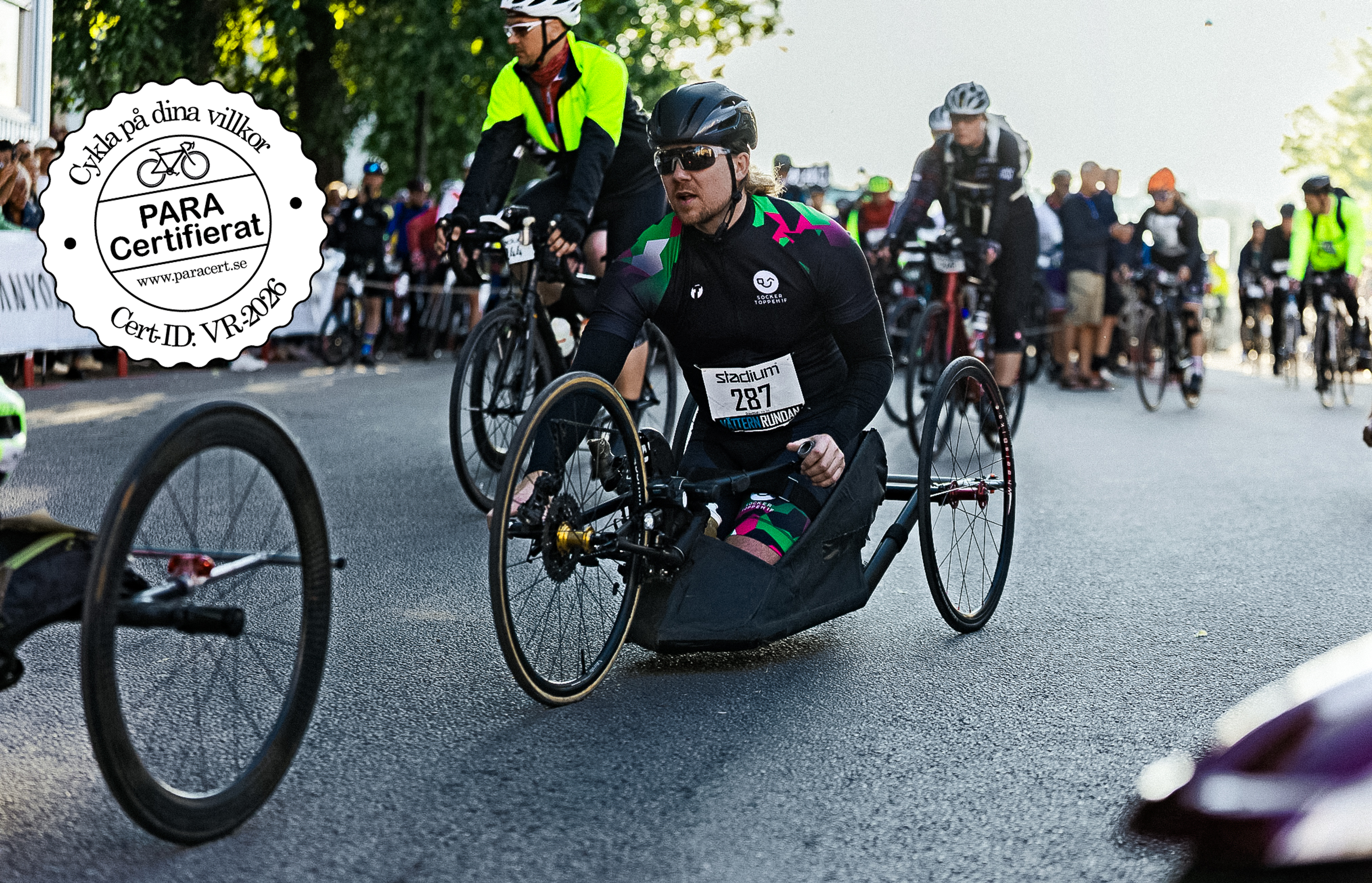 A handcyclist races on a road with other cyclists, with a "PARA Certifierat" logo in the top left.