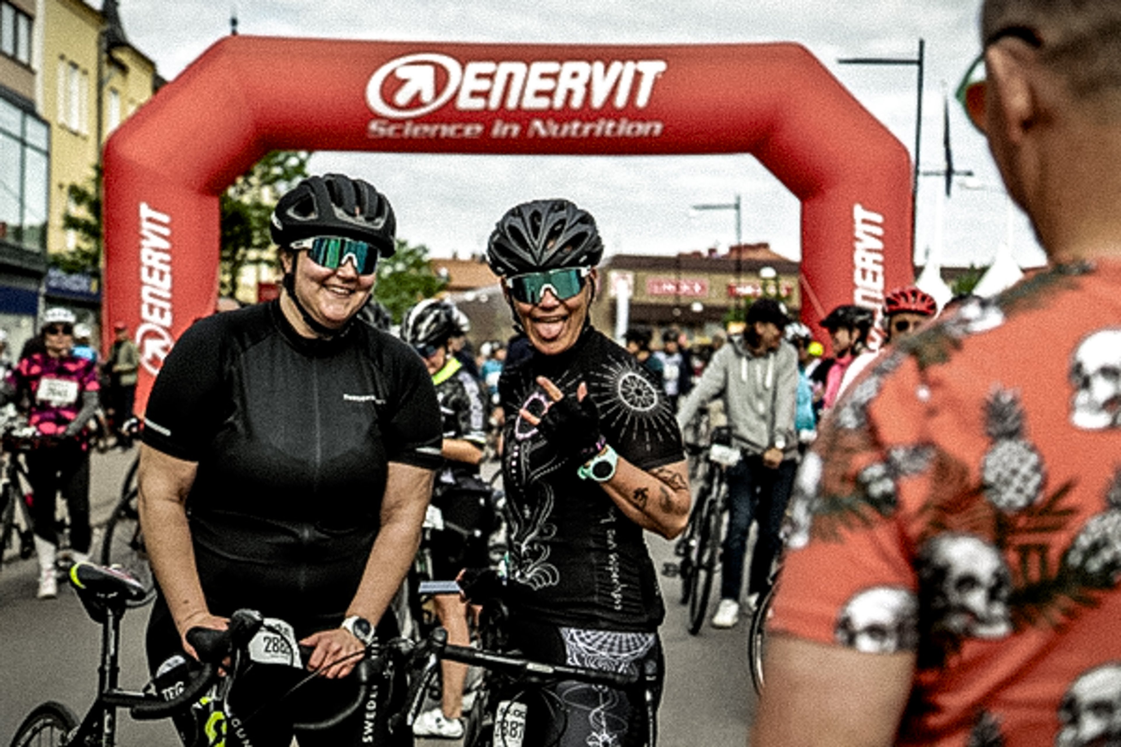 Two smiling cyclists, one with her tongue out, pose with their bikes under a red "ENERVIT" arch at an outdoor race event, with other participants in the background.
