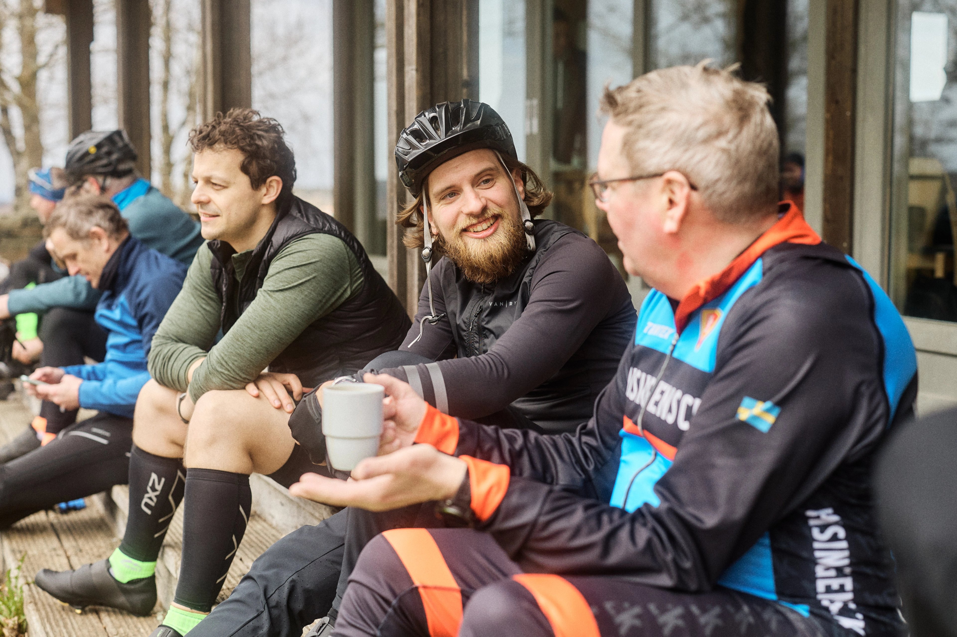 A group of cyclists resting outdoors, two men in the foreground smiling and conversing, one holding a cup.