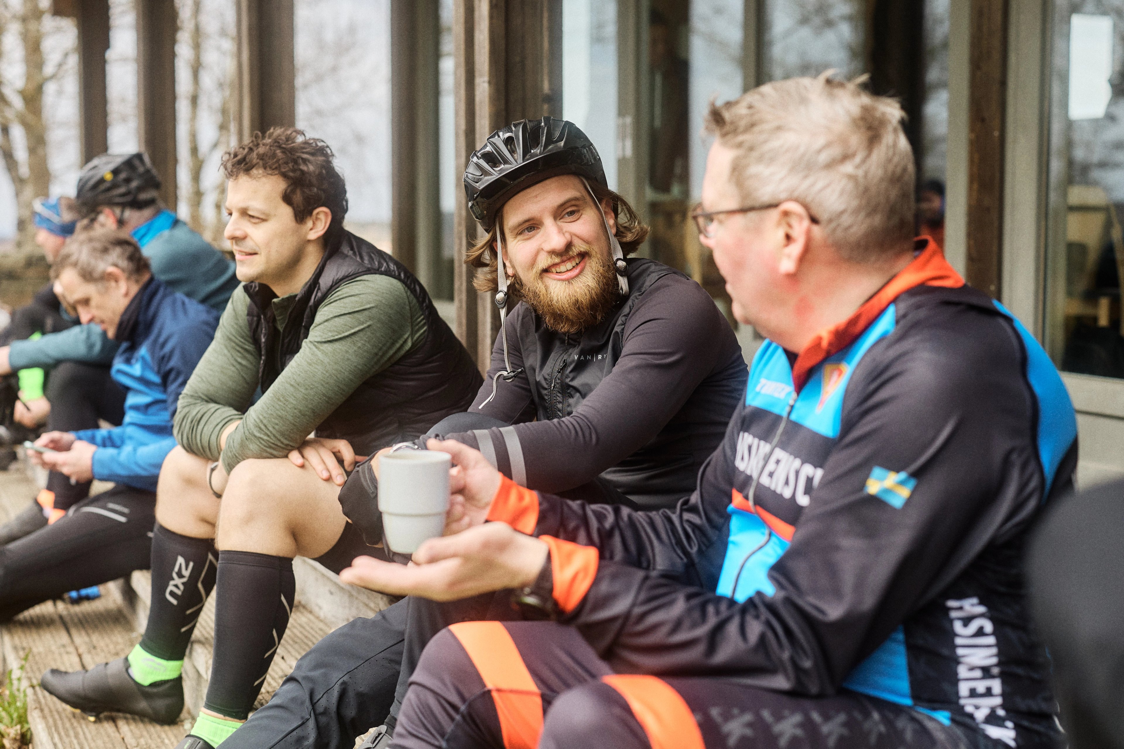 A group of cyclists resting outdoors, two men in the foreground smiling and conversing, one holding a cup.