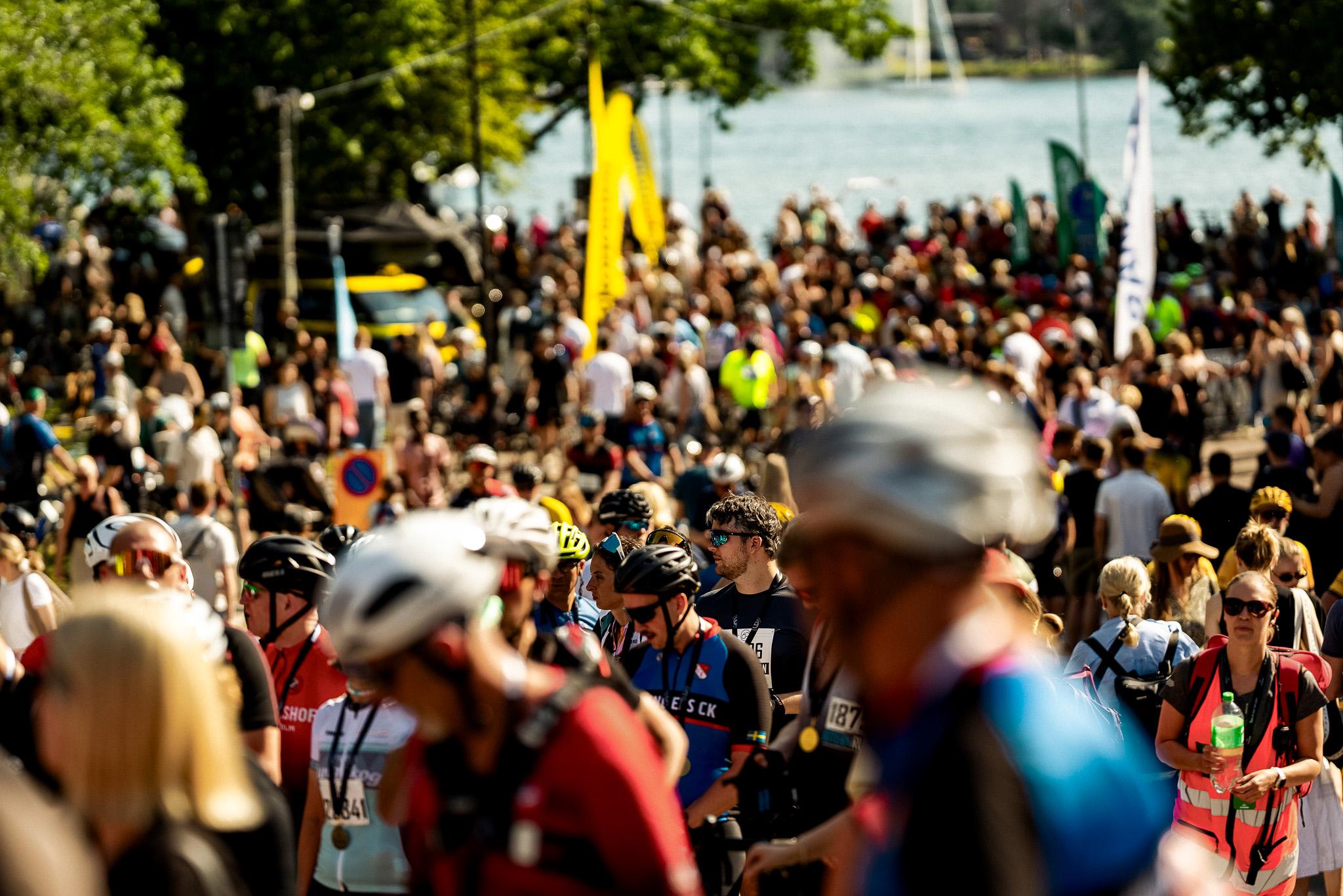 A large crowd of people, many in athletic gear and helmets, at an outdoor event near a body of water with flags.