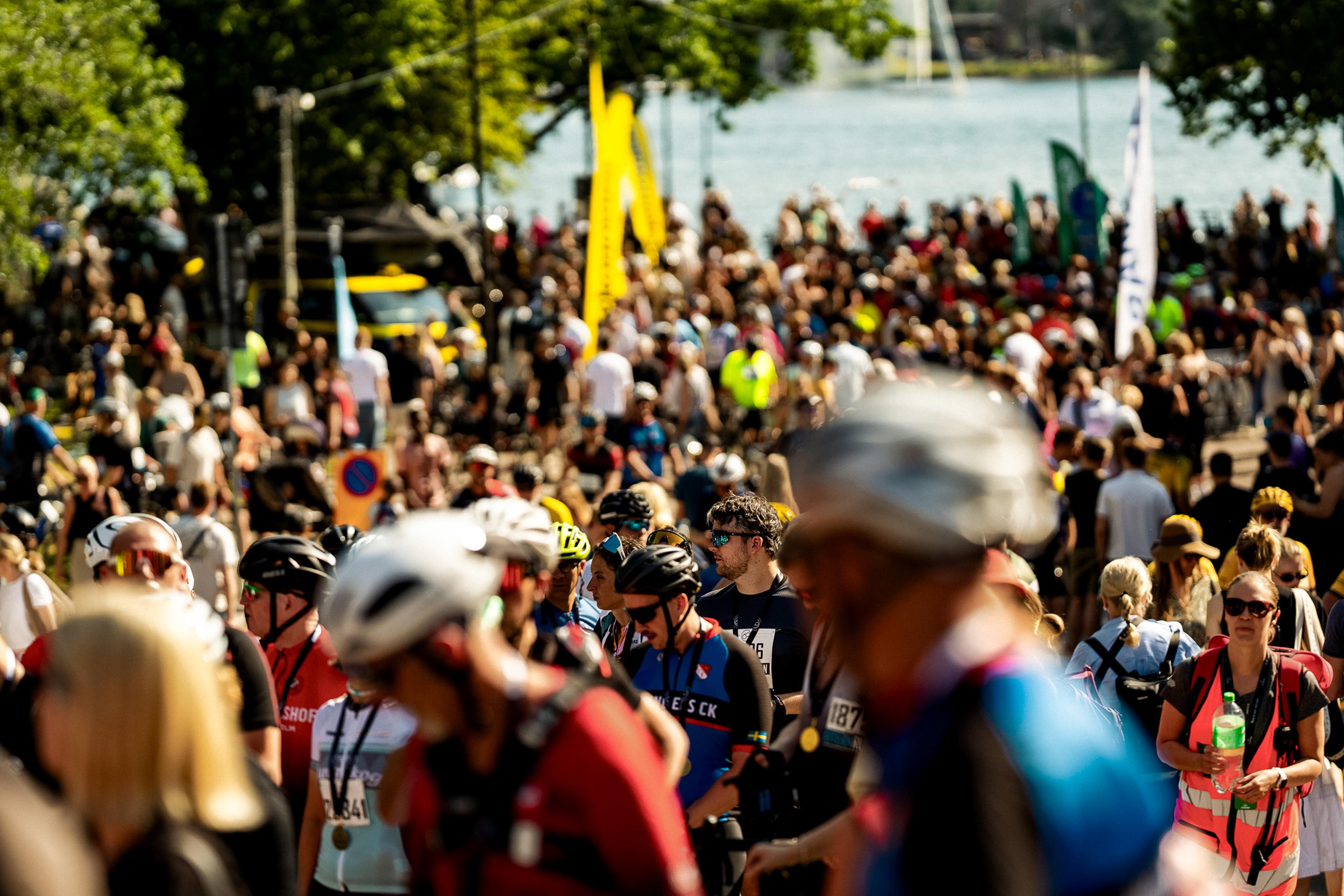 A large crowd of people, many in athletic gear and helmets, at an outdoor event near a body of water with flags.