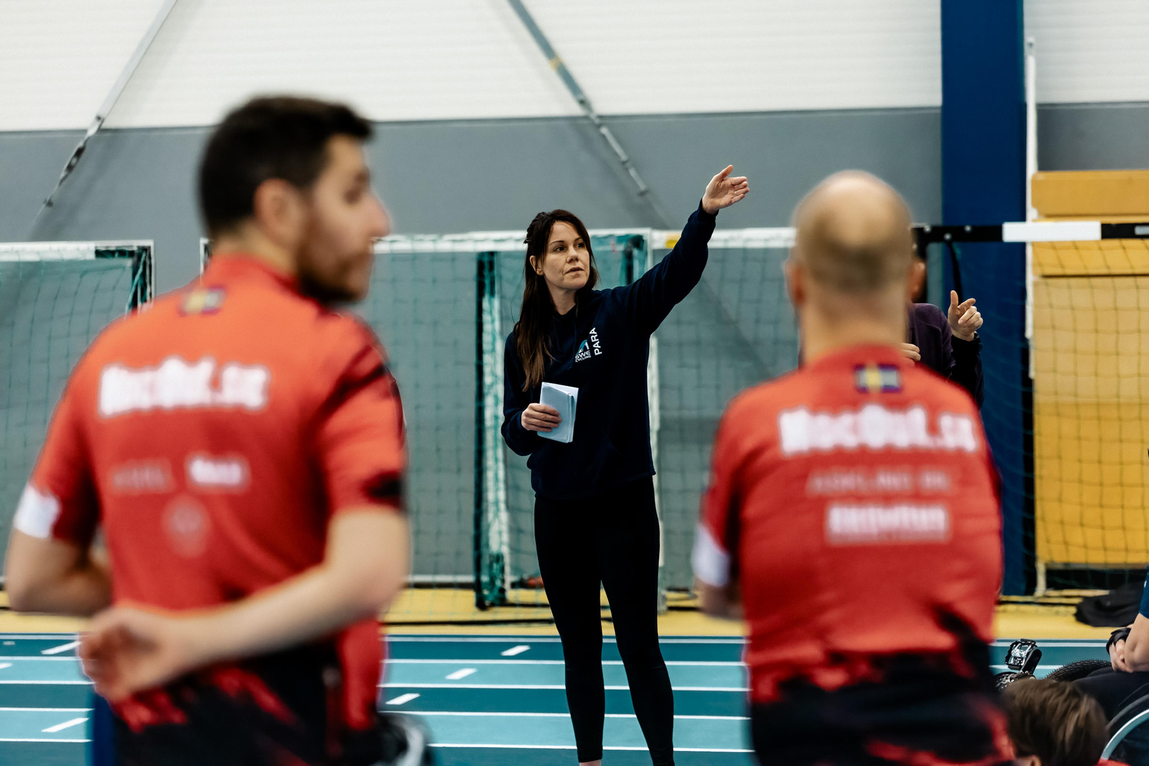 A woman gestures while talking to two athletes in red jerseys in an indoor sports hall.