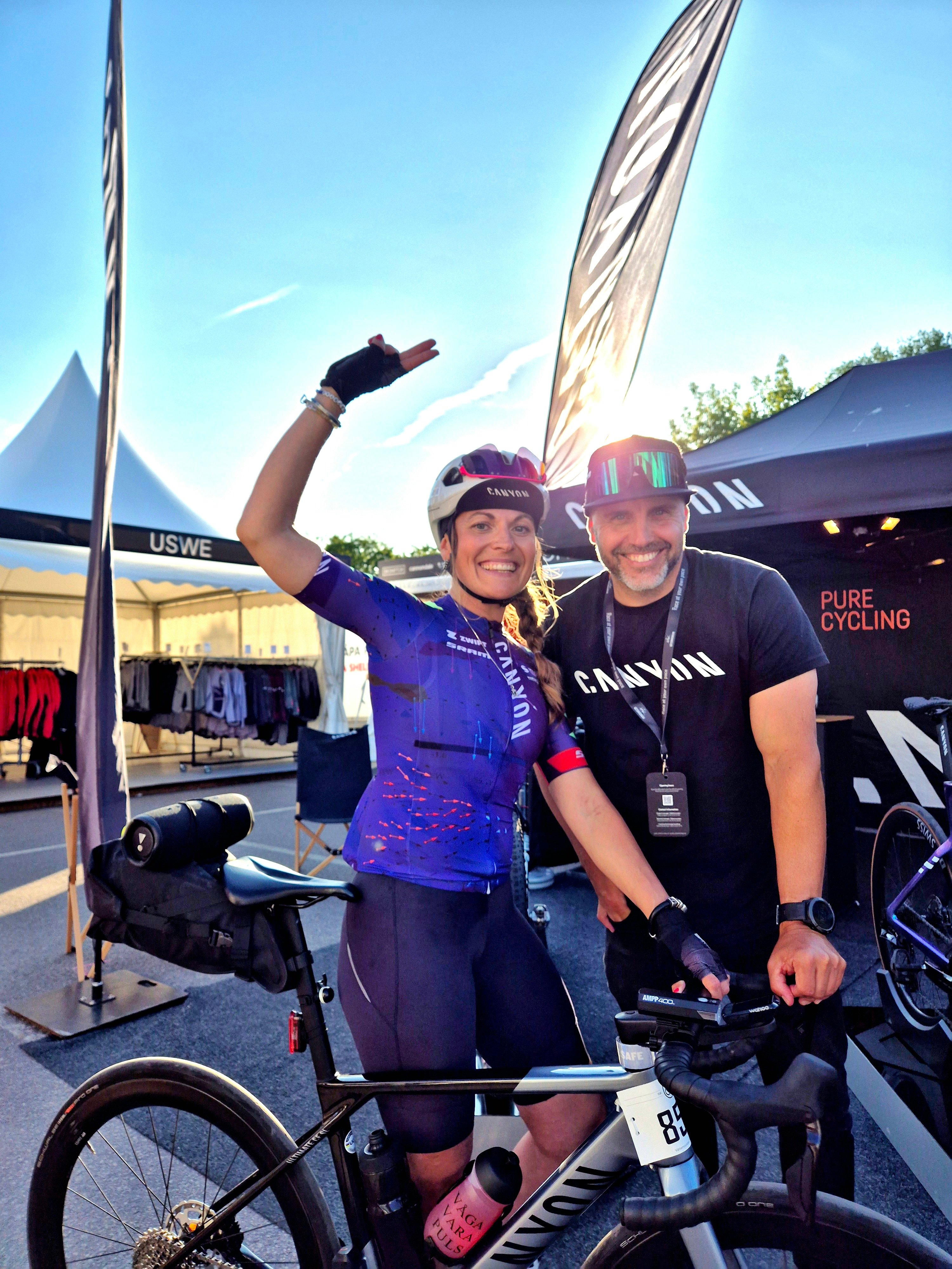 A smiling female cyclist in a purple jersey and a man in a Canyon shirt pose by a Canyon road bike at an outdoor event.
