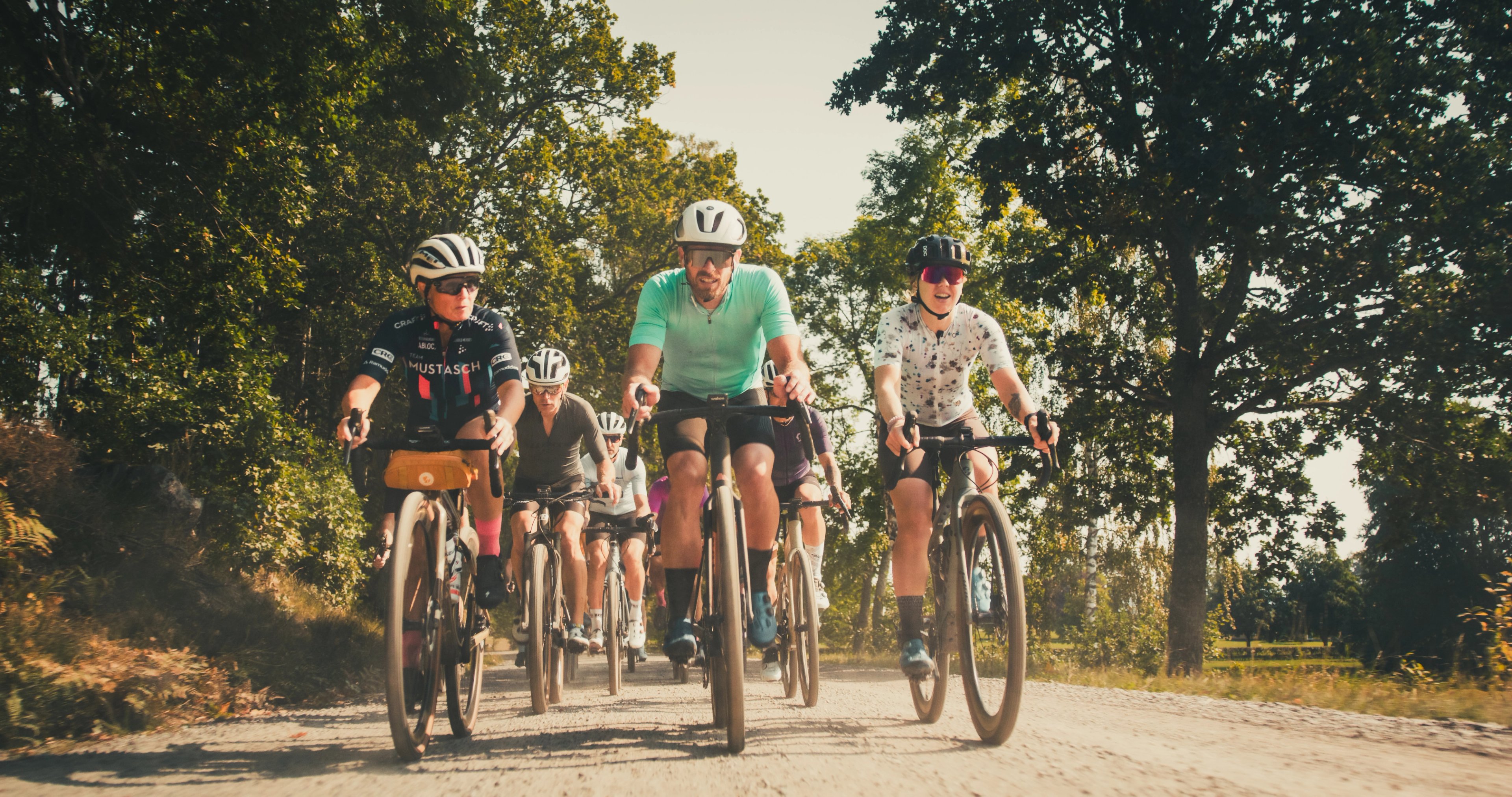 A group of cyclists rides gravel bikes on a sunlit dirt road lined with trees.
