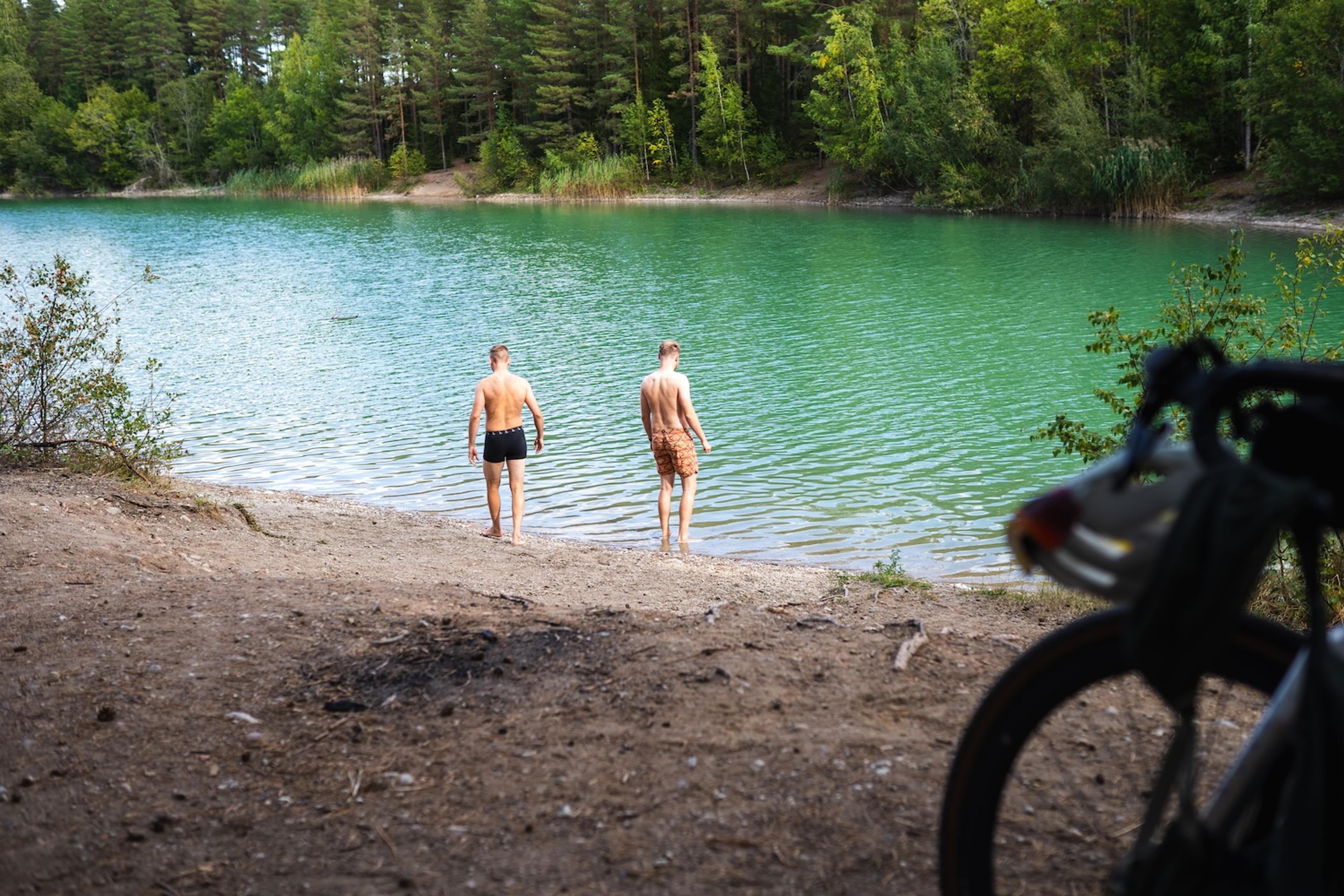 Two men walk into a turquoise lake from a dirt shore, with a bicycle in the foreground.