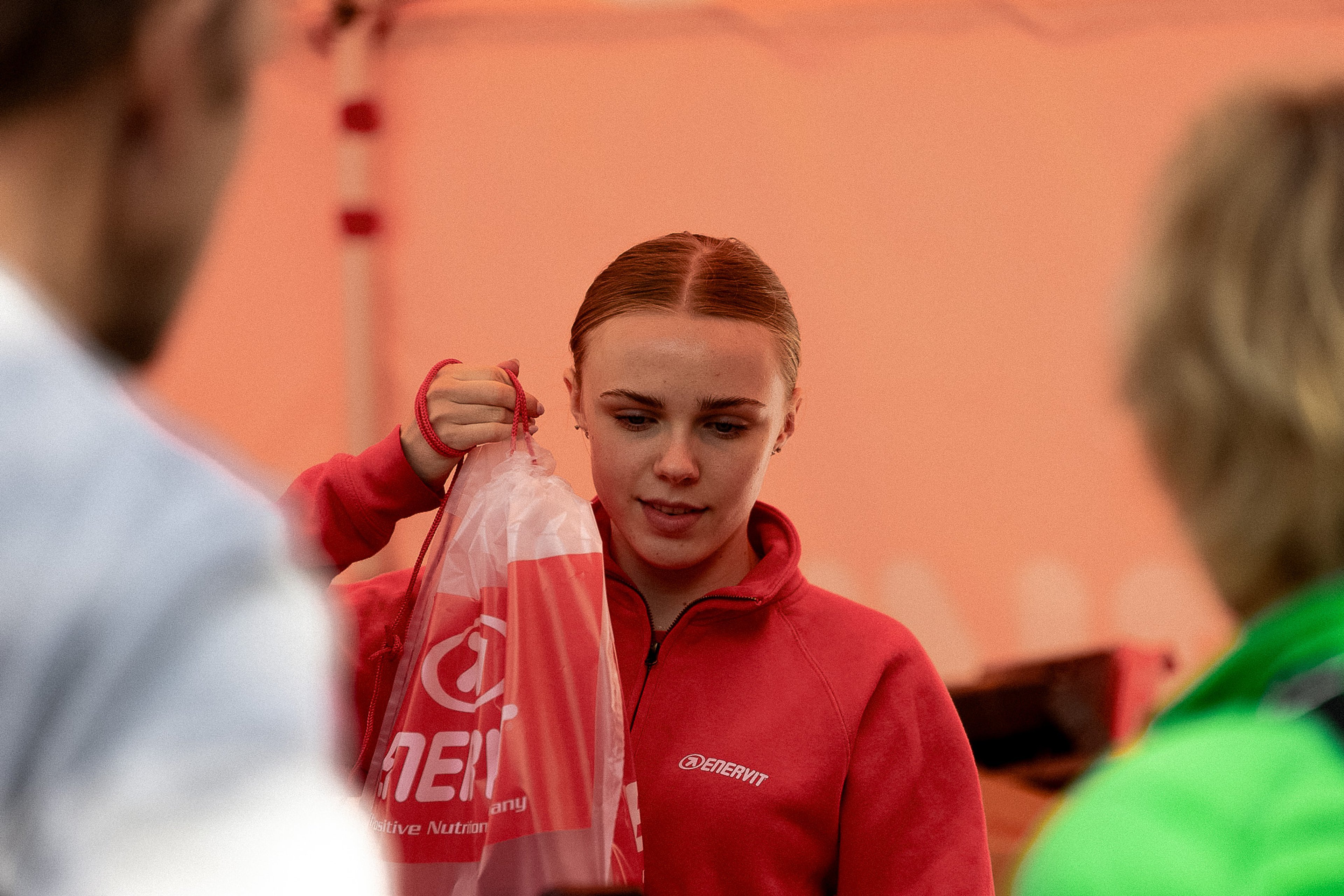 Young woman in a red Enervit jacket holding a branded plastic bag.