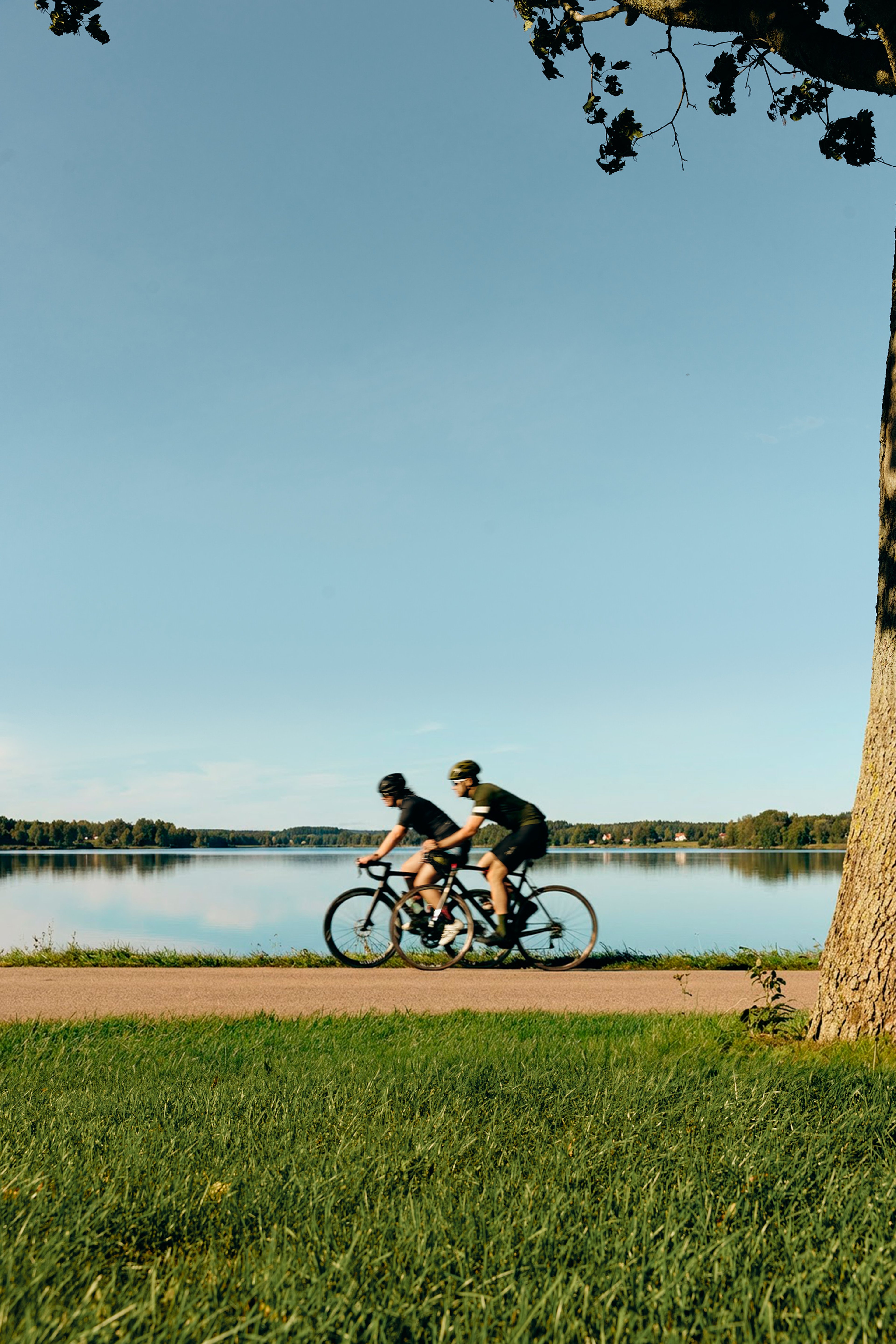 Two cyclists ride on a paved path next to a calm lake.