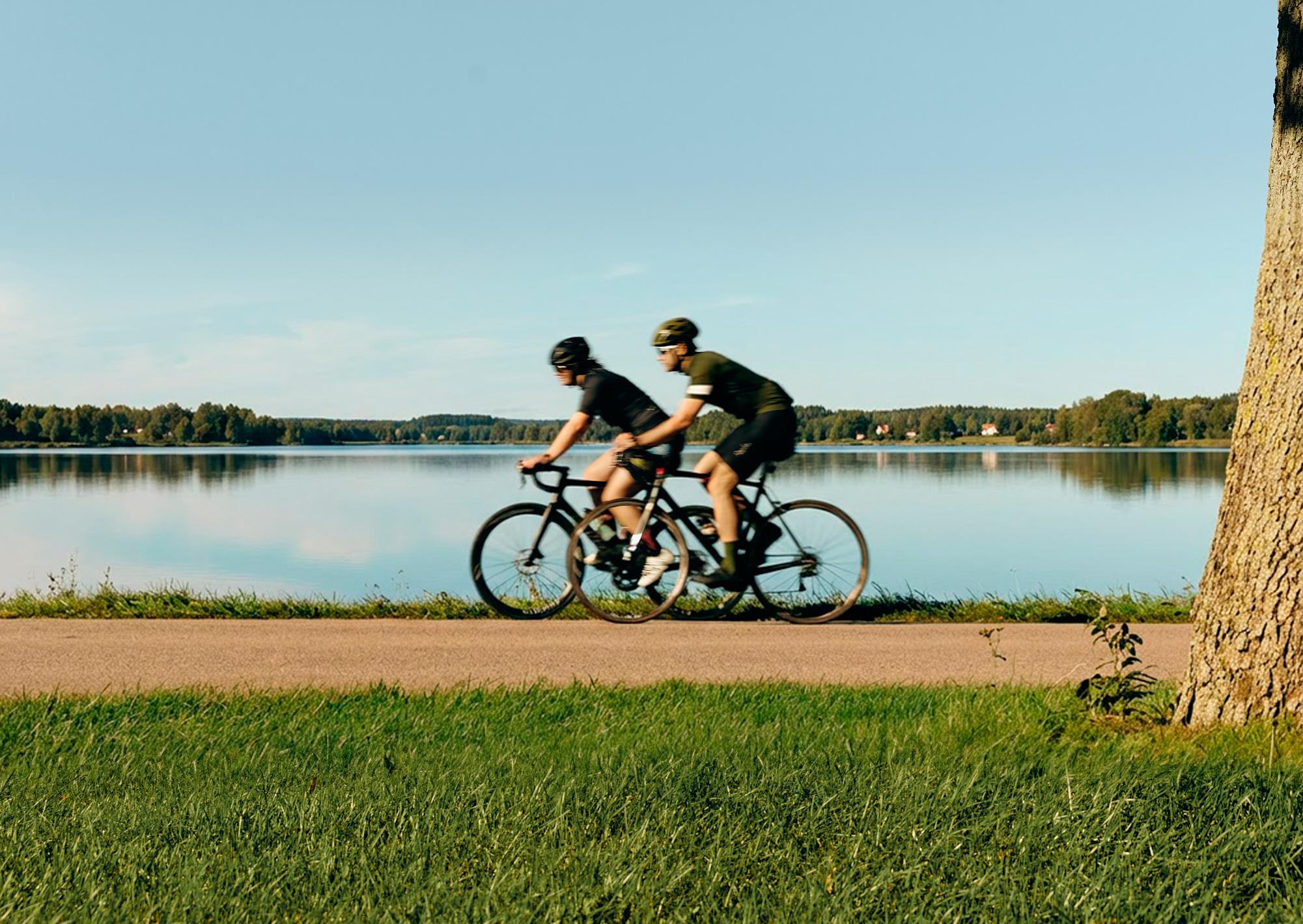 Two people ride bicycles on a path beside a calm lake under a clear blue sky.
