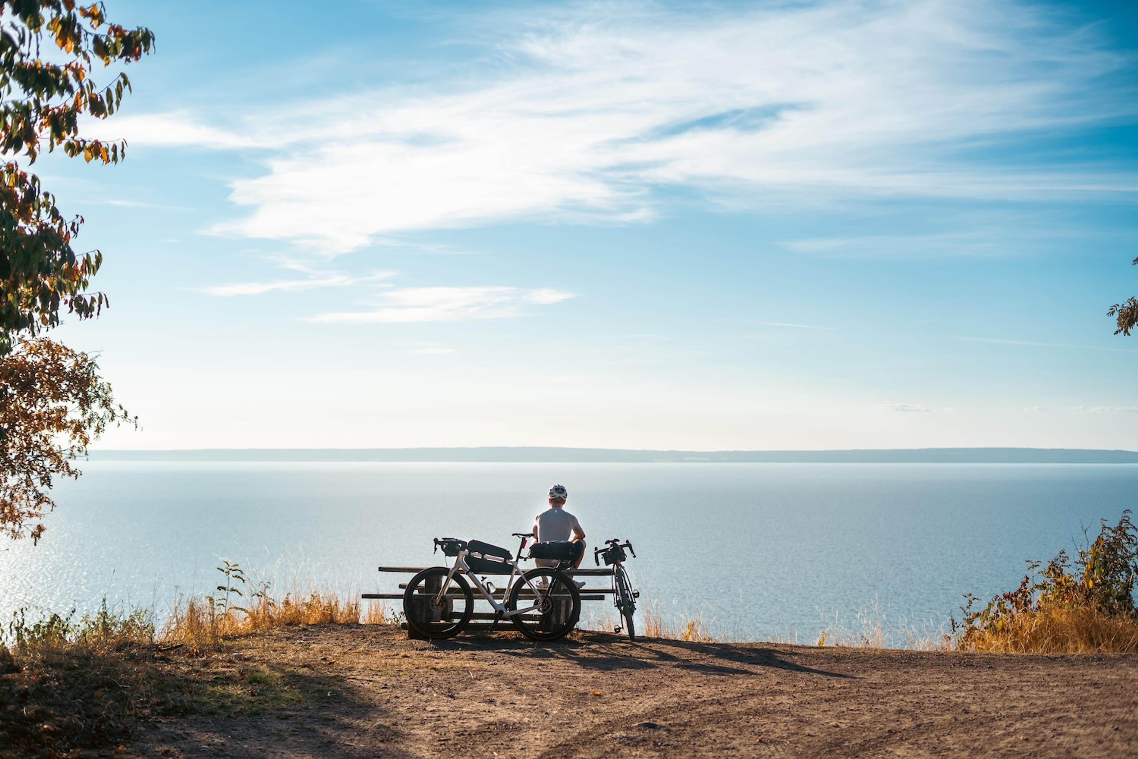 A person and their touring bike on a bench overlooking a calm lake under a blue sky.