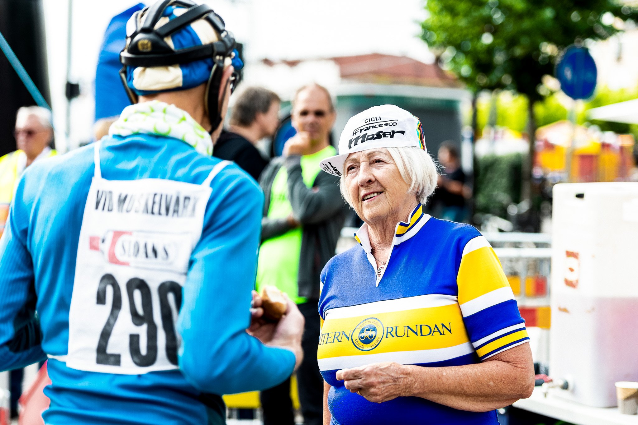 An older woman in a blue and yellow cycling jersey and white cap smiles while looking at a cyclist with race number 290 seen from behind.