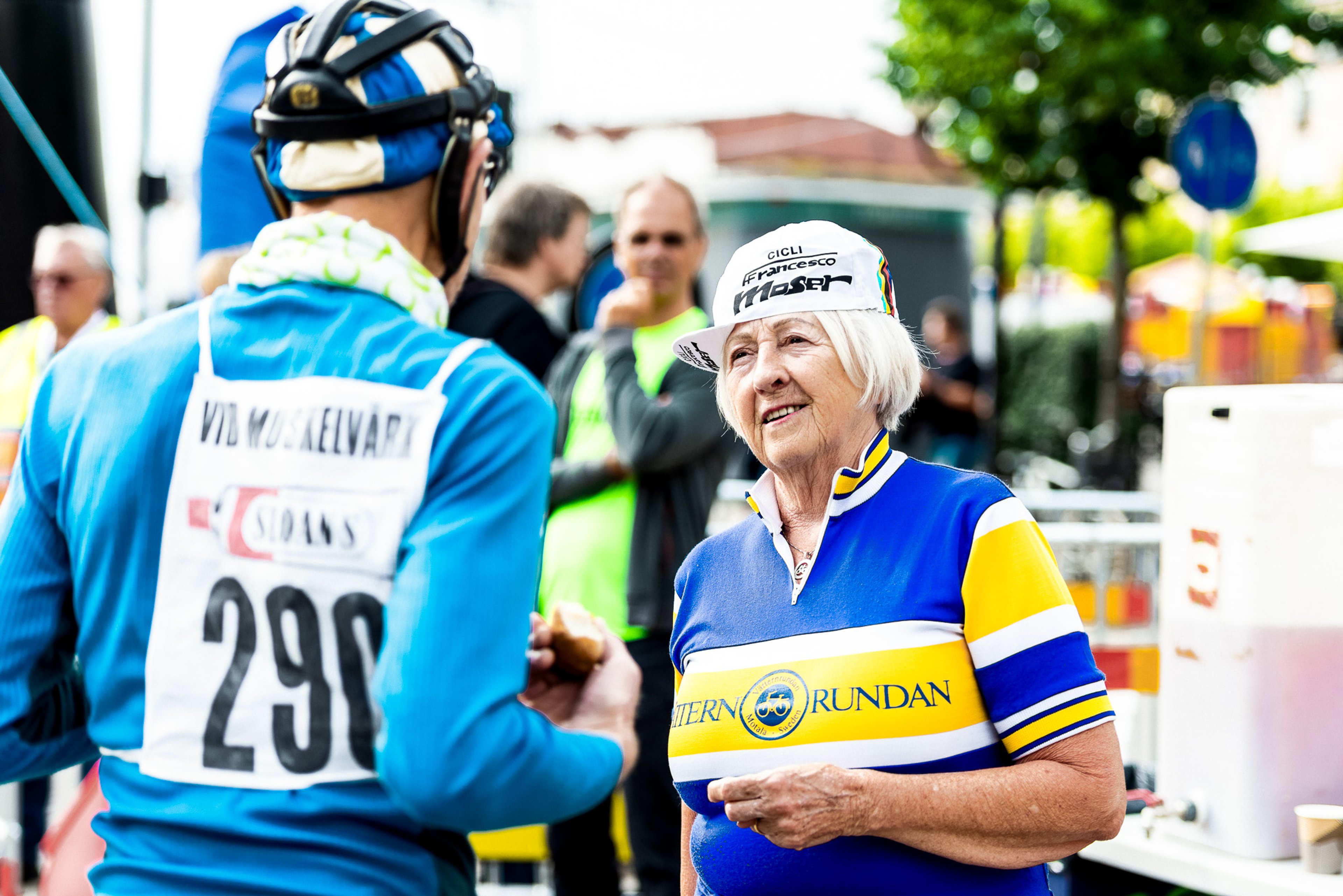 An older woman in a blue and yellow cycling jersey and white cap smiles while looking at a cyclist with race number 290 seen from behind.