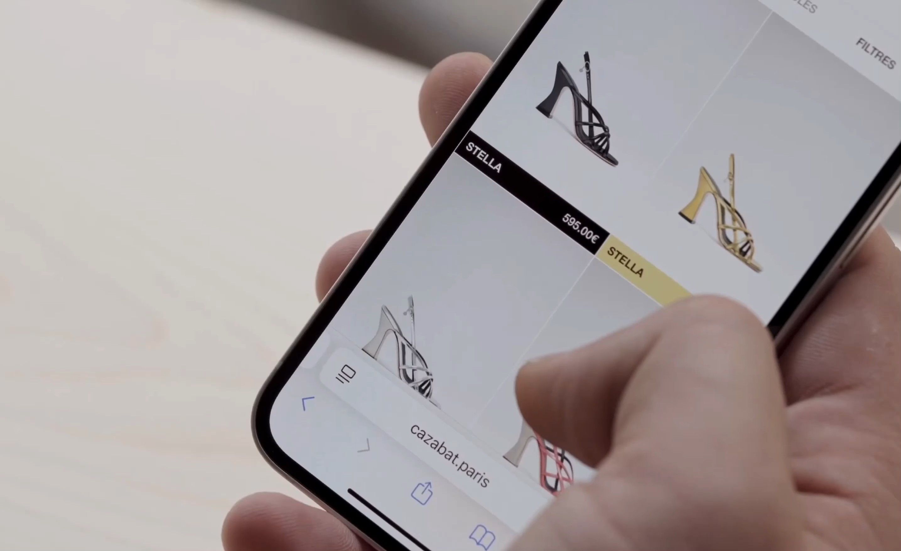 Hand holding an iPhone on a pale wood surface, opened to Cazabat’s Stella product page with the heel set against a yellow swatch.