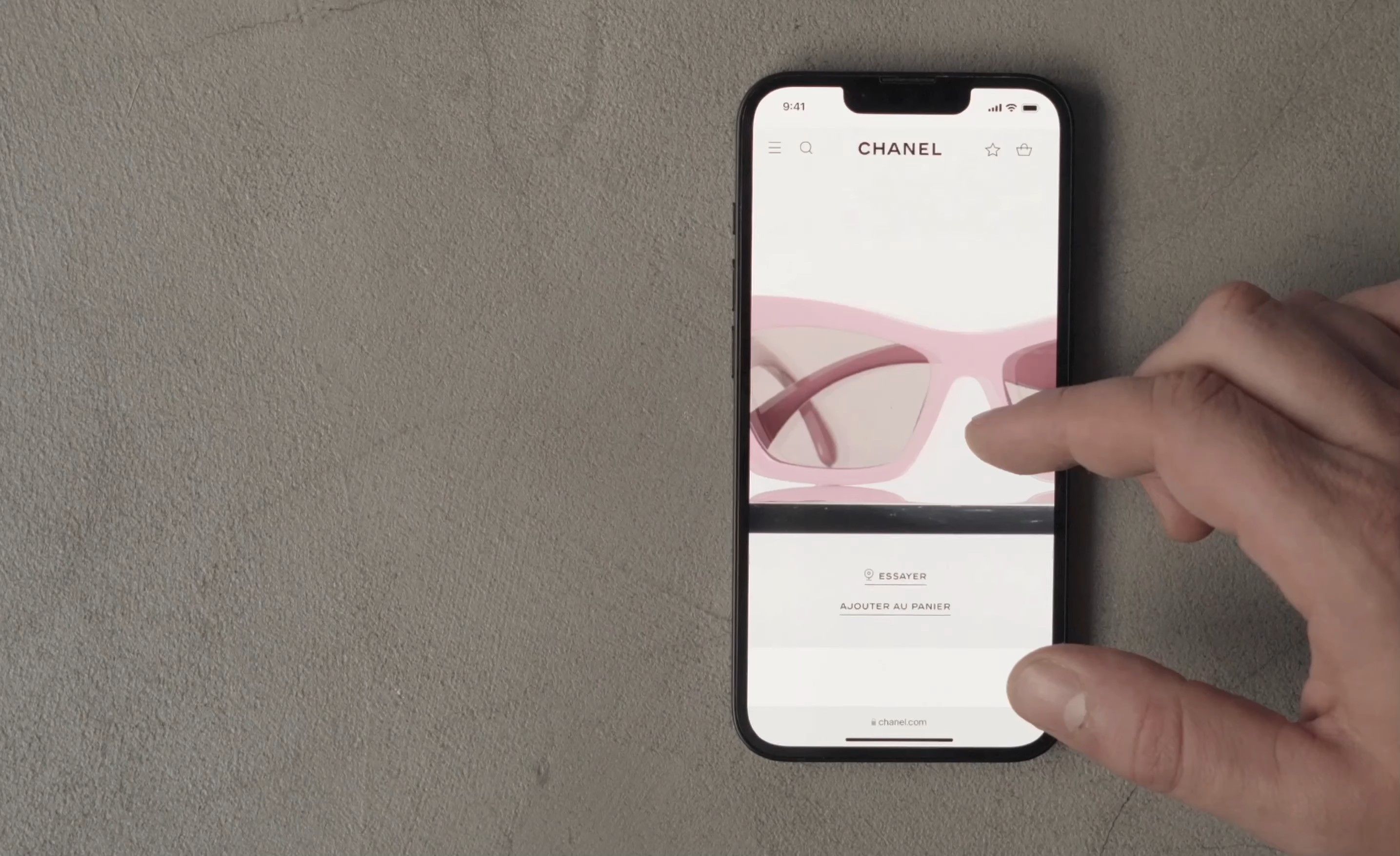 Hand holding an iPhone on a textured grey surface, displaying the CHANEL Eyewear site on a pink sunglasses product page.