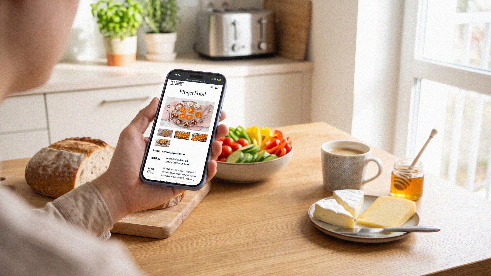 A hand holding a phone with the Catering Równość site; a breakfast table in the background