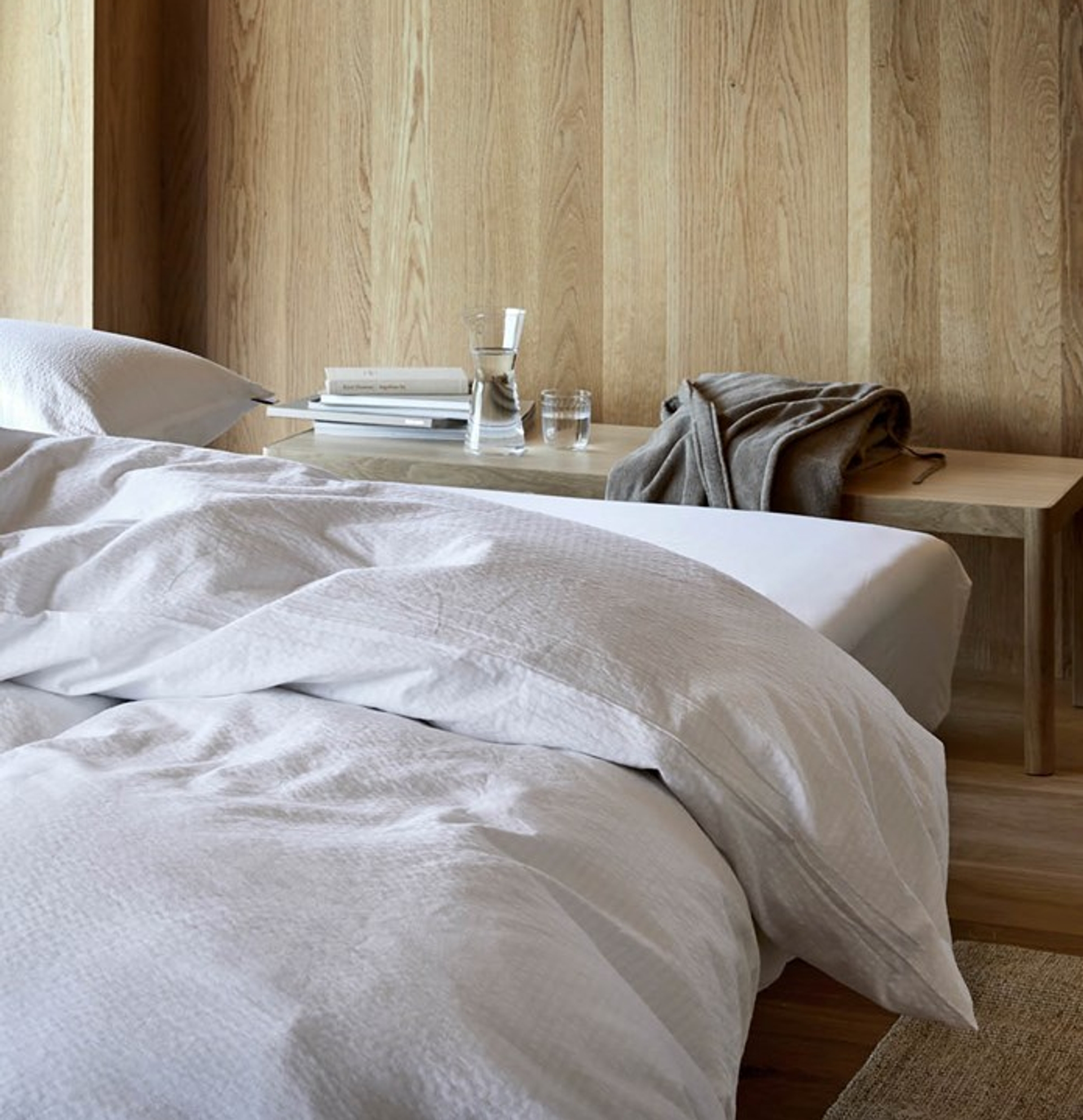 Alt-tekst: Elegant og minimalistisk soverom med hvitt kreppsengetøy lagt over en bred seng. Bakgrunnen består av vakre, naturlige trepaneler som gir rommet en varm og harmonisk atmosfære. Ved siden av sengen står en lav, lys trebenk med bøker, en vannkaraffel, et glass, og en grå morgenkåpe som er lagt over kanten. Gulvet er dekket av lyst treverk med et diskret vevd teppe som kompletterer den naturlige stilen.
