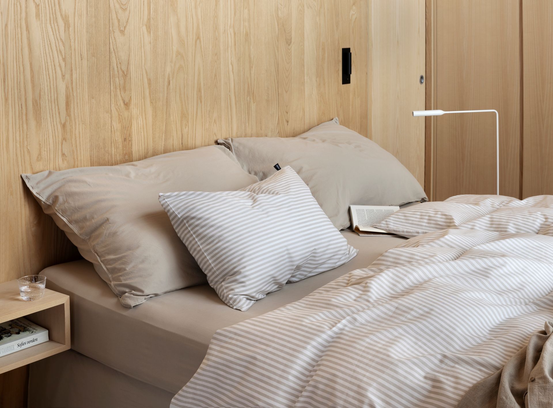 Detalj av oppredd seng med Høie Tor sengesett i beige – smale striper i myk bomull kombinert med ensfarget hotellputetrekk og laken, stylet med naturlige materialer og duse fargetoner for en rolig og enkel soveromsstil.