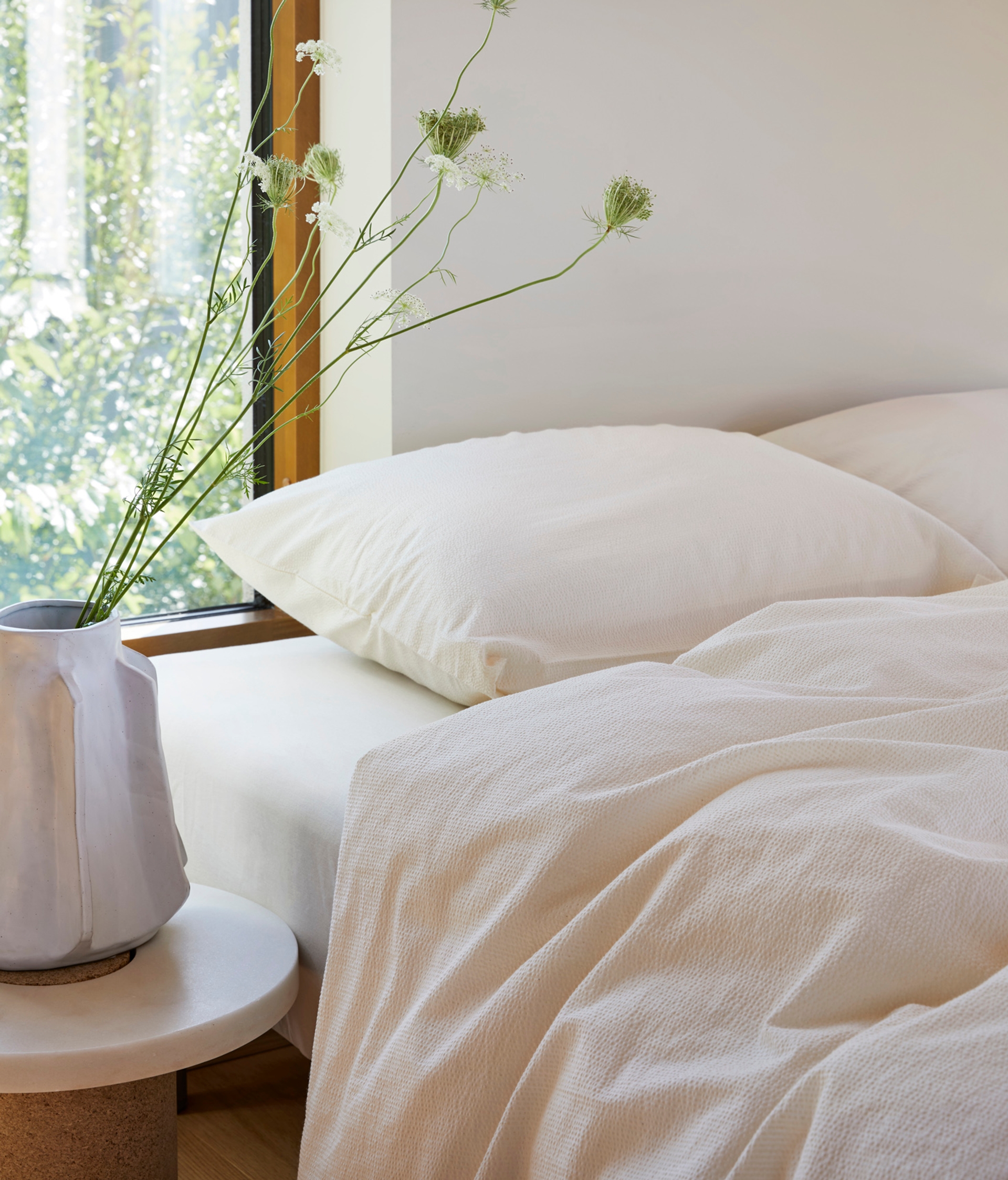 Lyst og minimalistisk soverom med ubleket kreppsengetøy som gir et rent og naturlig uttrykk. Sengen er plassert ved et stort vindu som slipper inn naturlig dagslys og gir utsikt til grønne omgivelser. På et lite, rundt bord står en hvit keramikkvase med lange, stilkete villblomster, som skaper en rolig og harmonisk atmosfære.