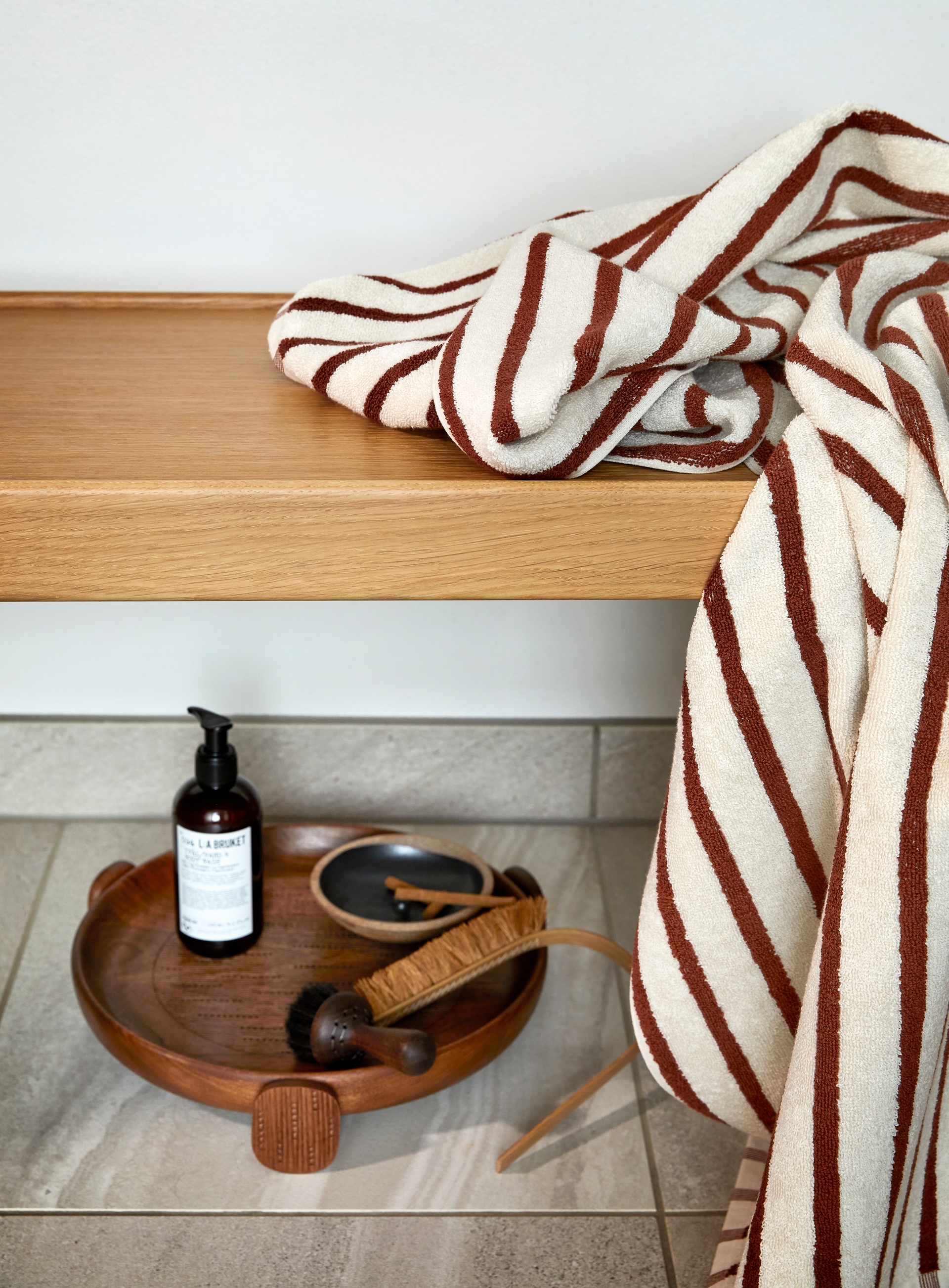 Et kremfarget håndkle med terrakottafargede striper ligger delvis brettet og drapert over en benk i lyst treverk. Under benken står et rundt brett i mørkt tre med velværeprodukter, inkludert en flaske såpe, en kroppsbørste og små skåler. Omgivelsene har et rolig og naturlig uttrykk med lyse fliser og myke kontraster.