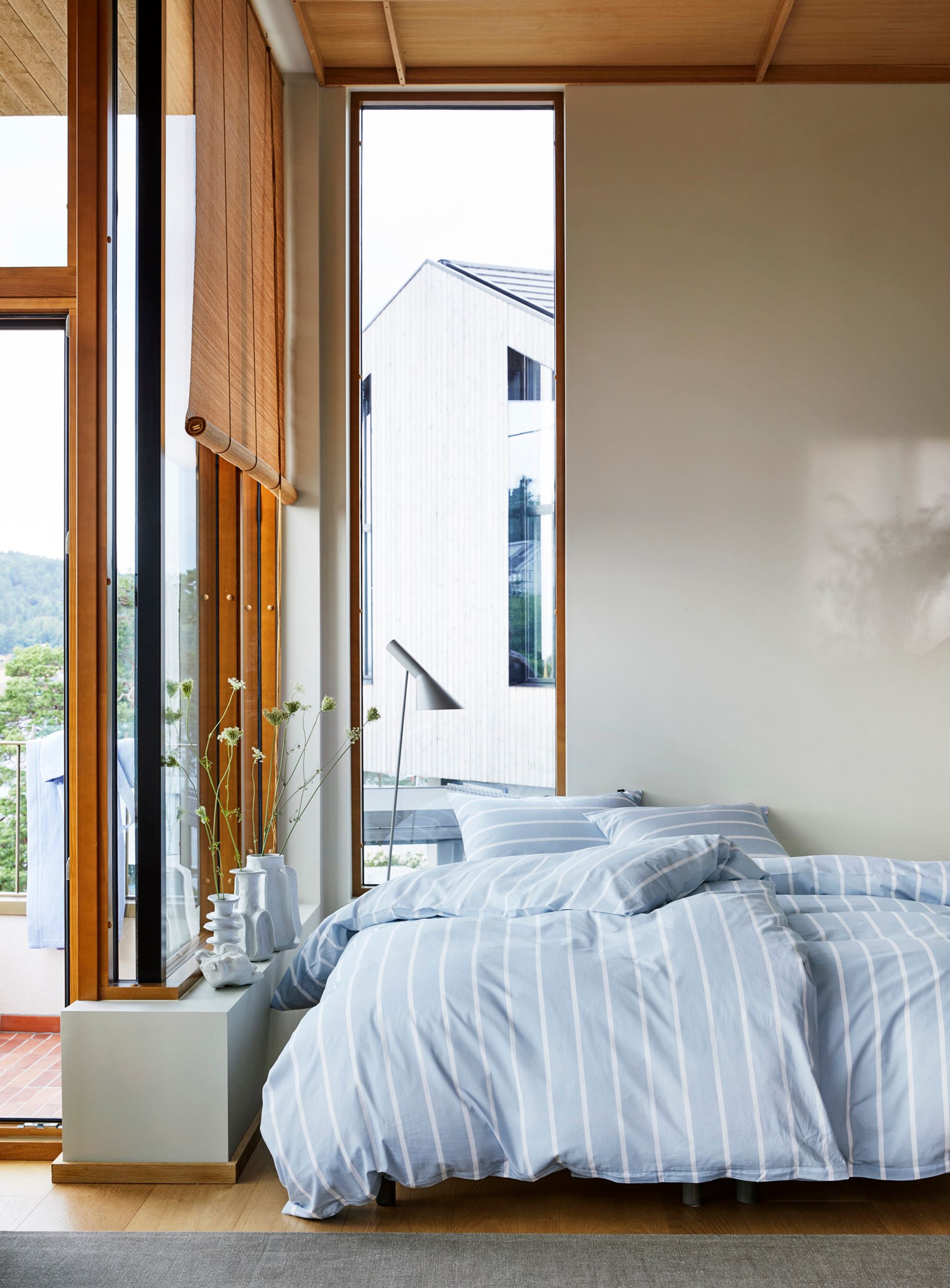 Høie Sander sengesett i denimblå og hvit stripet bomull – oppredd seng i et lyst og moderne rom med store vinduer og naturlig dagslys. Sengesettet har en frisk, tidløs stil med brede vertikale striper og en lett, luftig følelse.