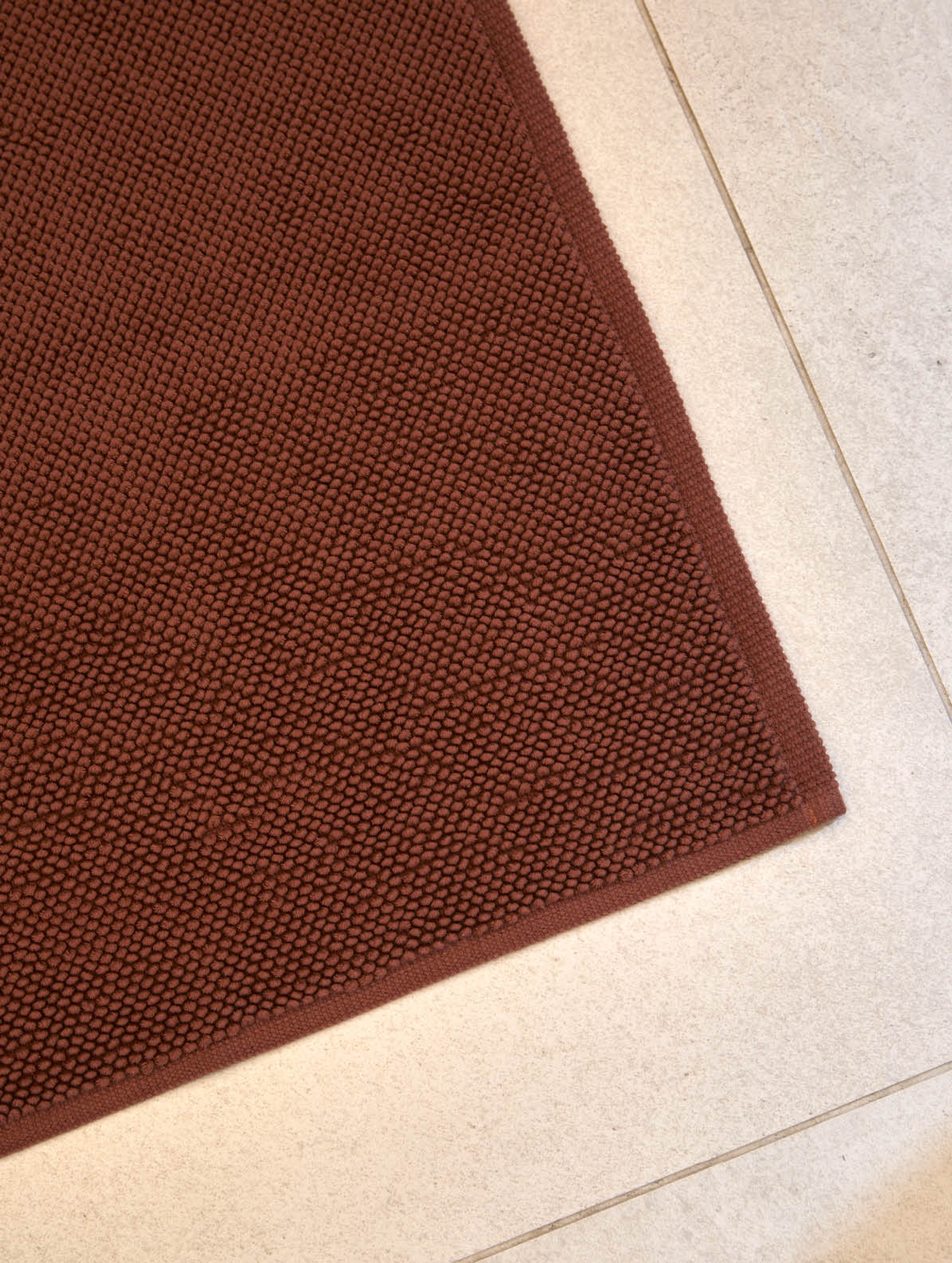 Nærbilde av en badematte i dyp terrakottafarge med strukturert overflate i små, jevne løkker. Matten ligger på et lyst flislagt baderomsgulv og har en innrammet kant som gir et rent og definert uttrykk.