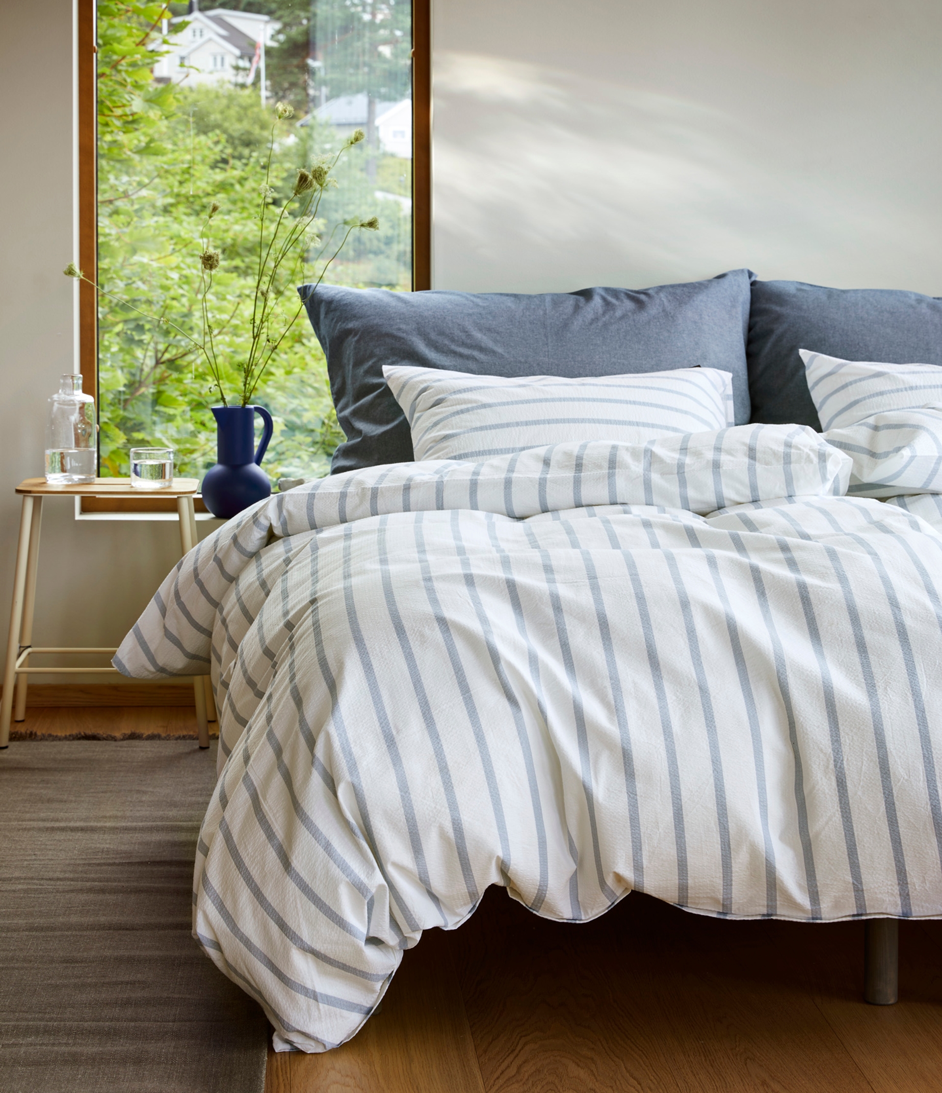 Oppredd seng med Høie Eilert sengesett i blå og hvit krepp – klassiske striper i kombinasjon med denimblå hotellputer gir et friskt, sommerlig uttrykk i et lyst soverom med utsikt til grønne omgivelser.