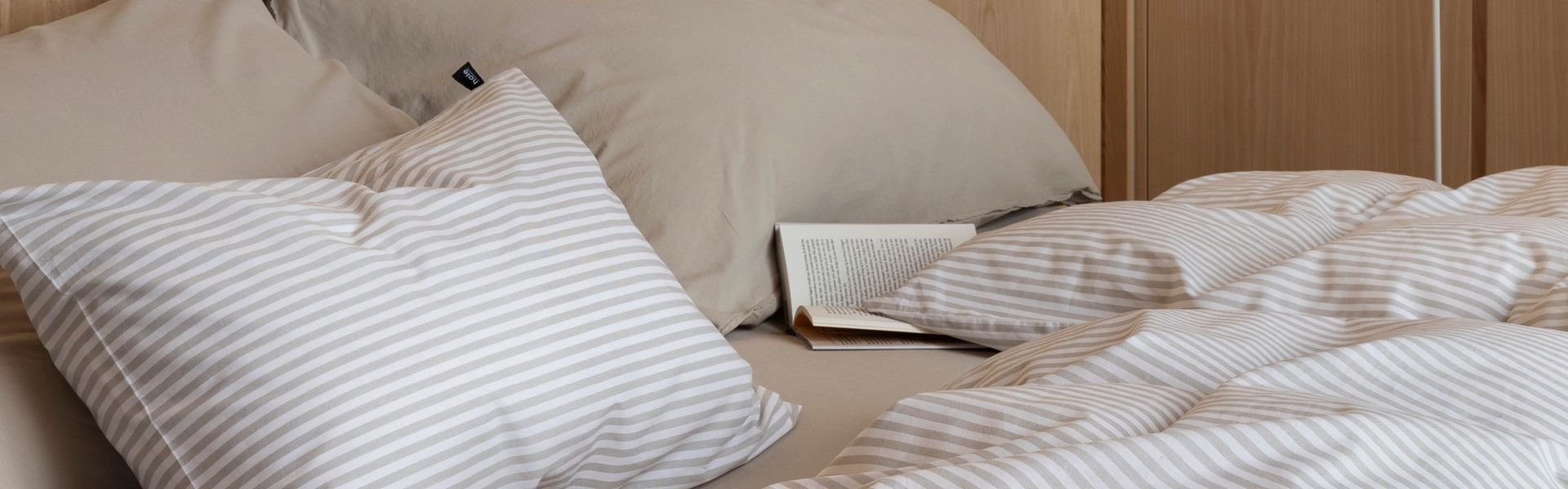 Detalj av oppredd seng med Høie Tor sengesett i beige – smale striper i myk bomull kombinert med ensfarget hotellputetrekk og laken, stylet med naturlige materialer og duse fargetoner for en rolig og enkel soveromsstil.