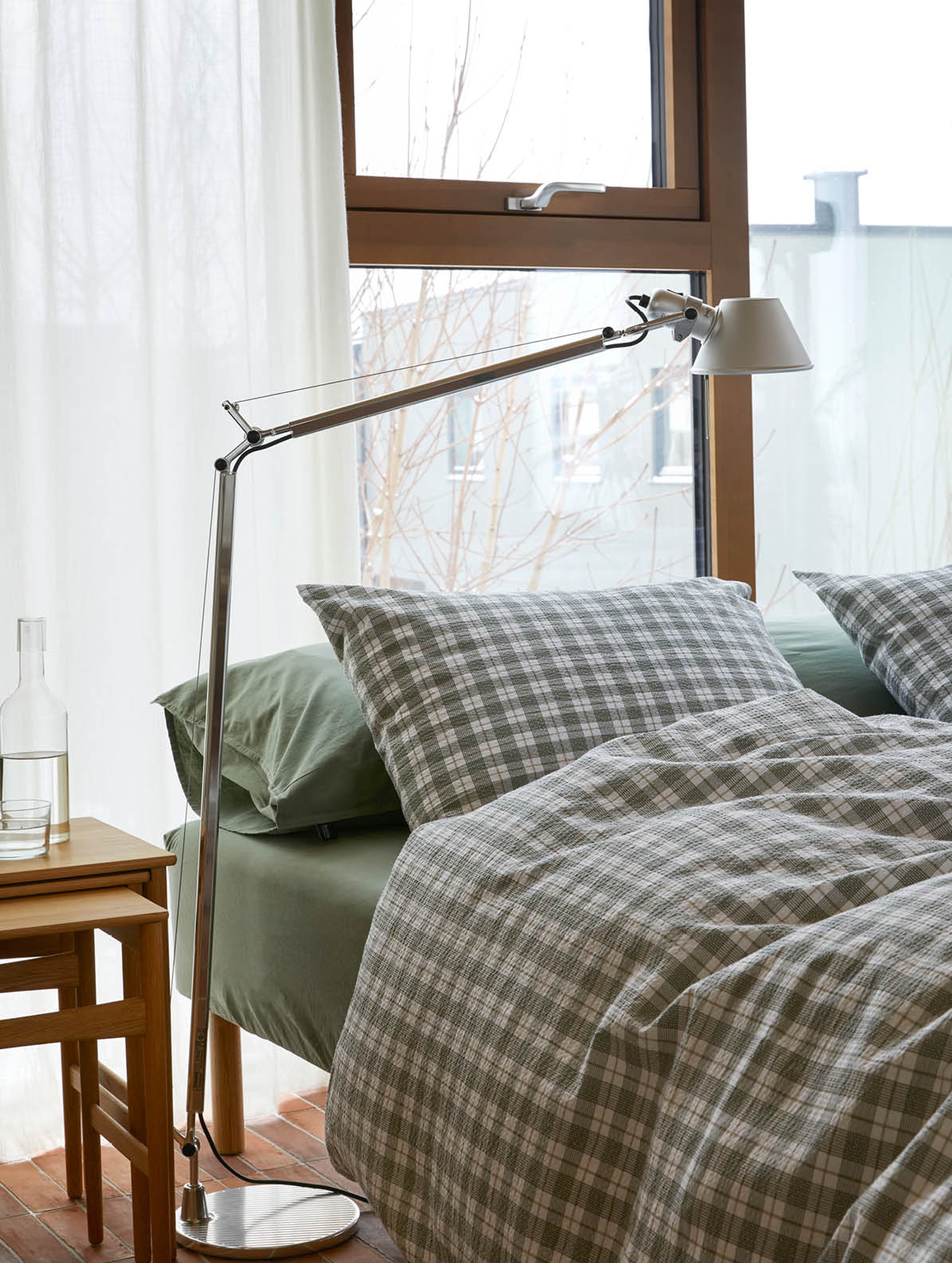 Soverom med moderne, industrielt preg med en betongvegg i bakgrunnen. Sengen er oppredd med rosa sengetøy og blomstrete kreppdyne i lyse nyanser av rosa, gult og grått, som skaper et mykt og feminint uttrykk. Ved siden av sengen står en hvit leselampe på et lite bord, omgitt av bøker og magasiner på gulvet.