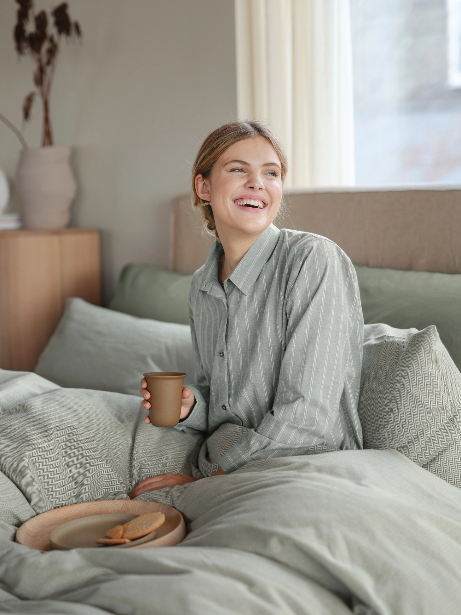 Kvinne som nyter frokost i sengen iført myk flanellpyjamas i dempet grønn med klassiske striper – komfortabel pysjamas i flanell for dame, perfekt til rolige morgener og hjemmekos.