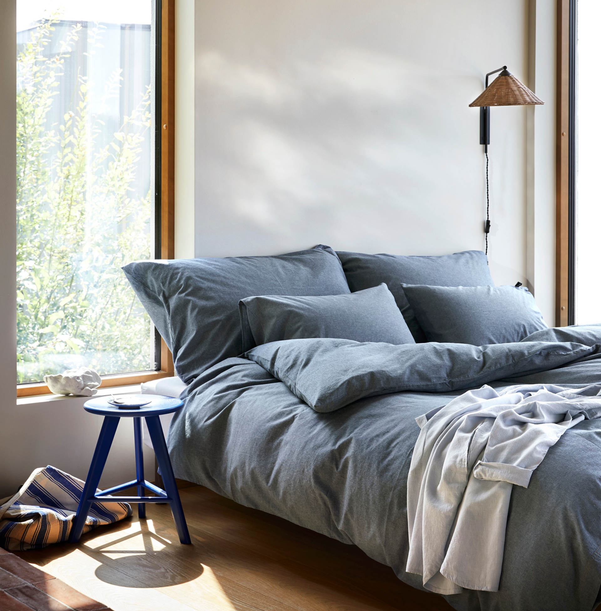 Oppredd seng med Høie Ola sengesett i garnfarget denimblå – laget av ekstra fin bomull for en myk og naturlig følelse, stylet i et lyst rom med store vinduer og minimalistiske detaljer.