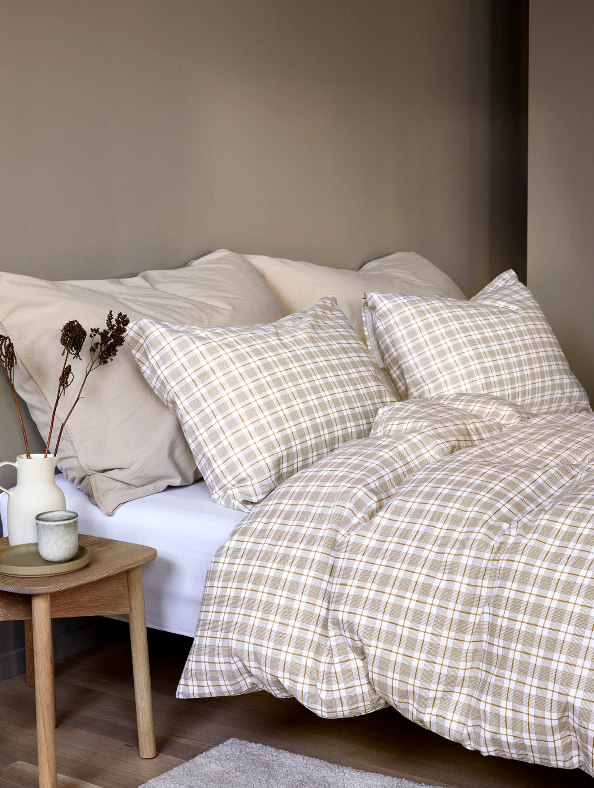 Lyst og luftig soverom med hvitt sengetøy dekorert med delikate, grønne bladmotiver som gir et friskt og naturlig preg. Sengen er delvis dekket av en lys grønn skjorte som tilfører et mykt fargeinnslag. Ved siden av sengen står en beige vase med høye, stilkete villblomster som forsterker det naturlige uttrykket. Store vinduer med hvite gardiner slipper inn rikelig med dagslys og gir utsikt til grønne omgivelser. En moderne stol med grønt sete står ved siden av sengen.