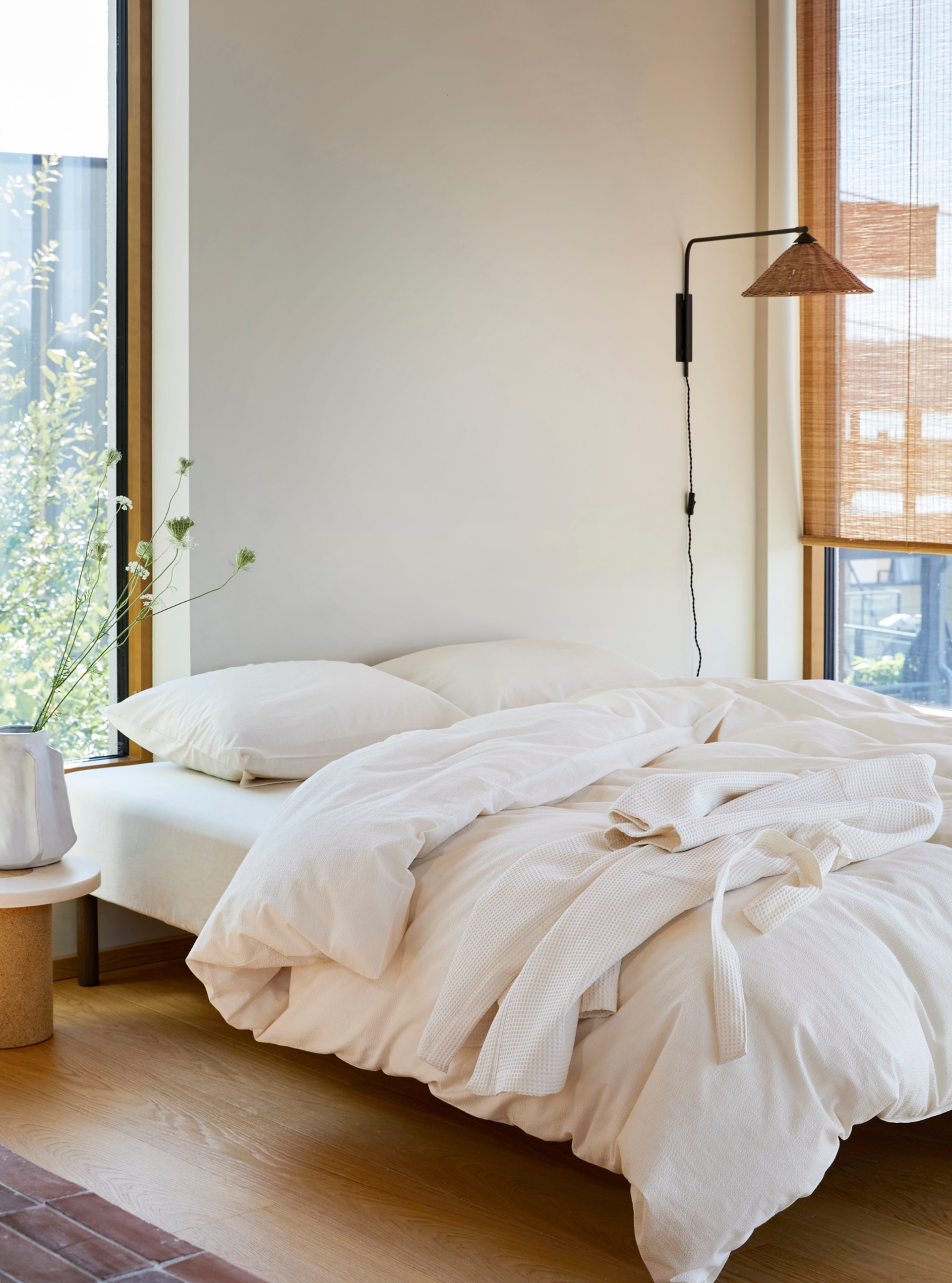 Alt-tekst: Lyst og minimalistisk soverom med ubleket kreppsengetøy og en ubleket morgenkåpe lagt over sengen. Sengen er plassert ved store vinduer som slipper inn naturlig dagslys og gir utsikt til grønne omgivelser. En vase med lange, stilkete villblomster står på et lite bord ved siden av sengen, mens en veggmontert lampe med naturlig rotting-skjerm skaper en lun og harmonisk atmosfære.