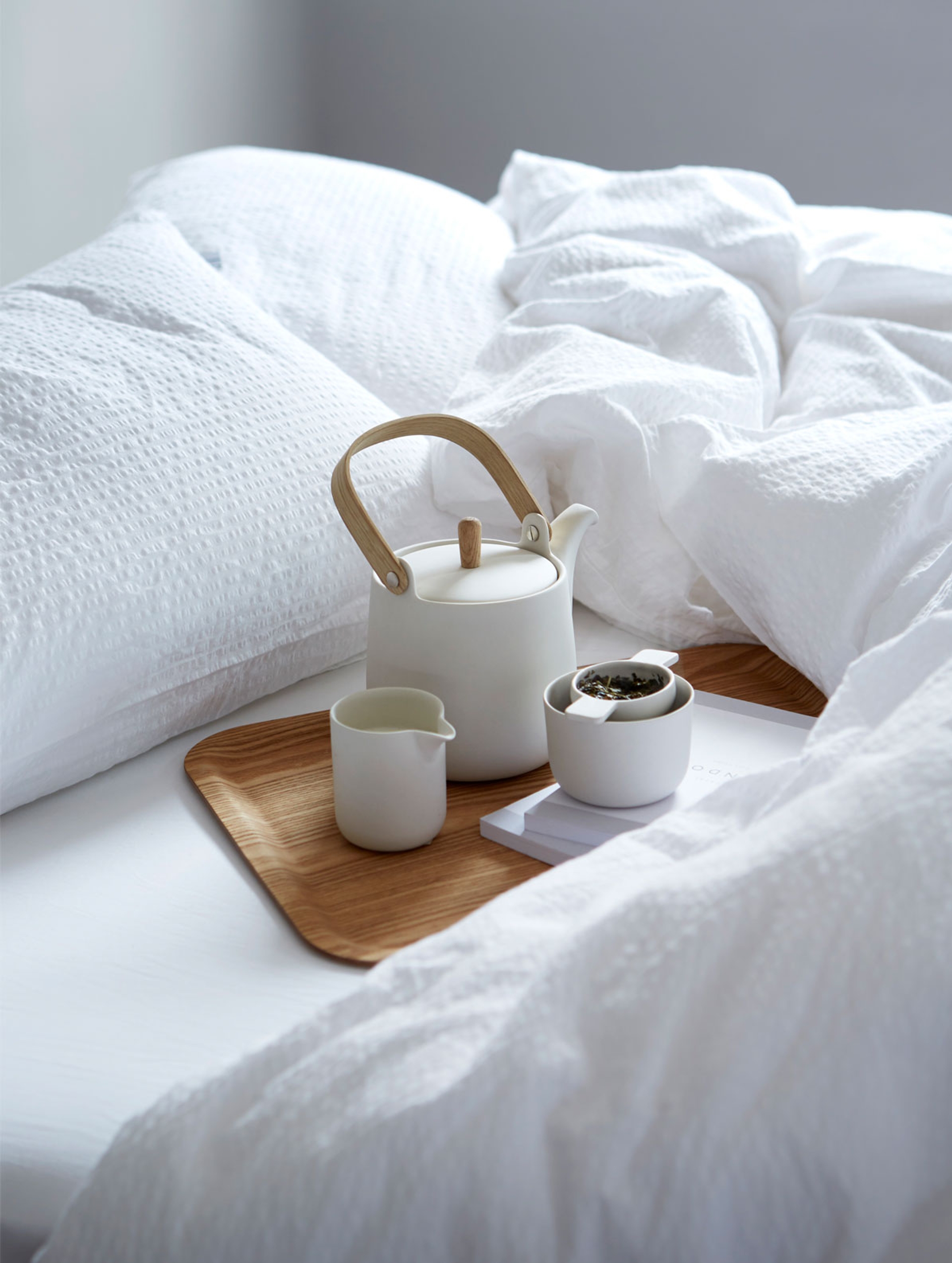 Hvit, luftig seng med kreppsengetøy som gir en ren og avslappende atmosfære. På sengen står et naturlig trefat med en moderne, hvit tekanne med håndtak i lyst tre, en matchende fløtemugge, og en liten bolle med teblader. Under fatet ligger en bok, som gir et inntrykk av en rolig morgenstund med te på sengen.