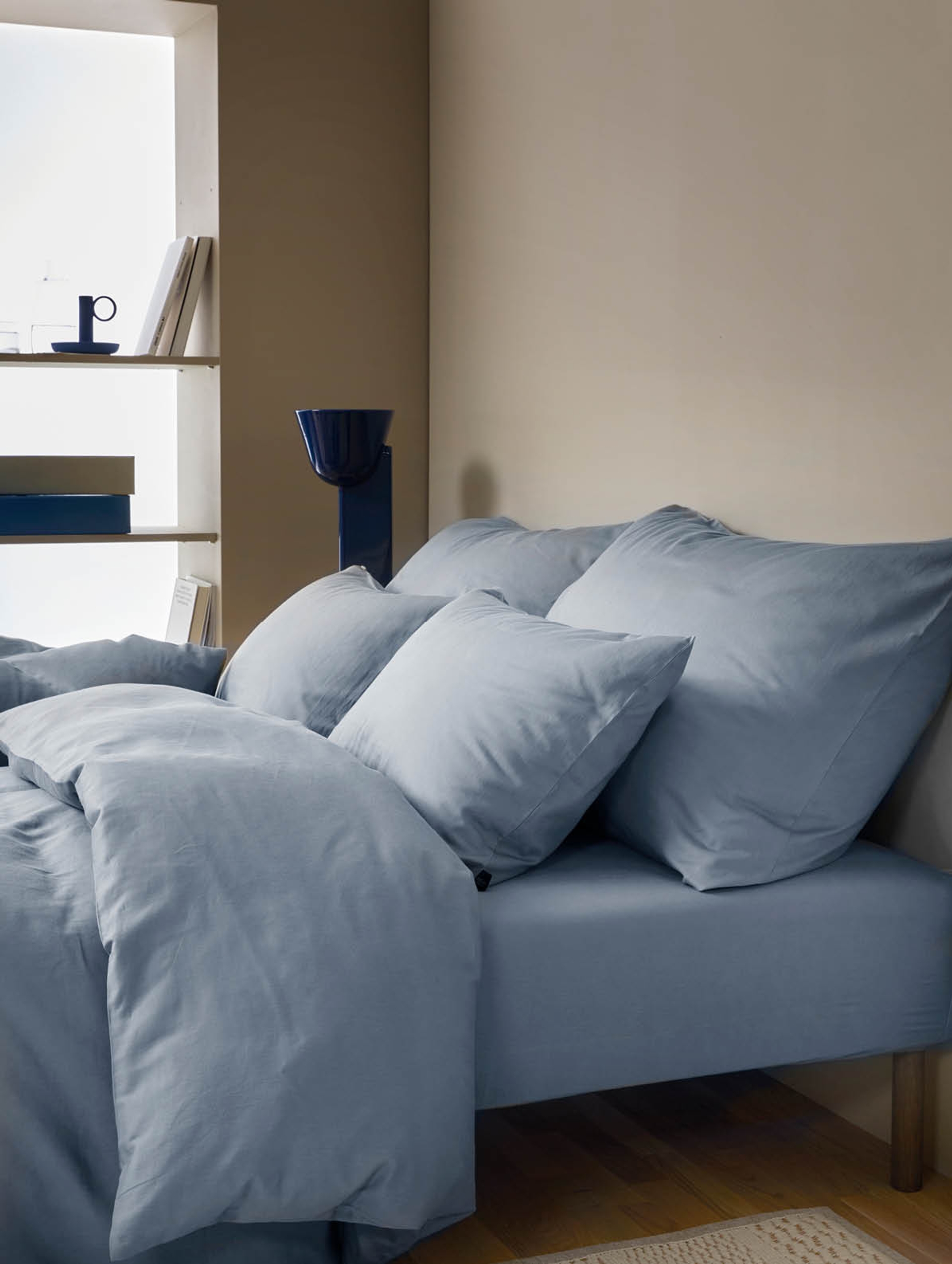 Dobbeltseng med harmonisk sengetøy i dus blå steinvasket sateng, pyntet med hotellputer og vanlige puter. Myke tekstiler mot en lun, beige vegg med minimalistisk interiør i bakgrunnen.