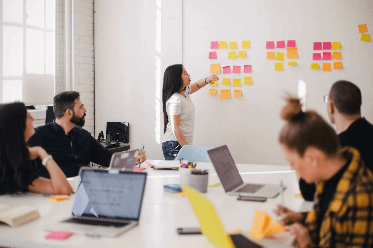 A team collaborates in a modern meeting room, using sticky notes on a wall and laptops on a table to brainstorm ideas.