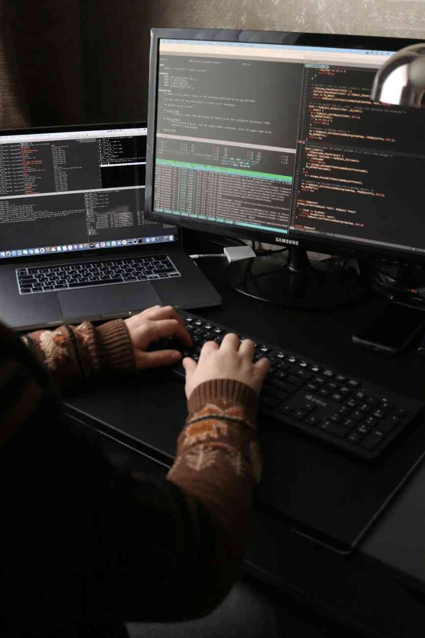 A person in a patterned sweater types on a keyboard at a desk with dual computer screens displaying code and terminal outputs.
