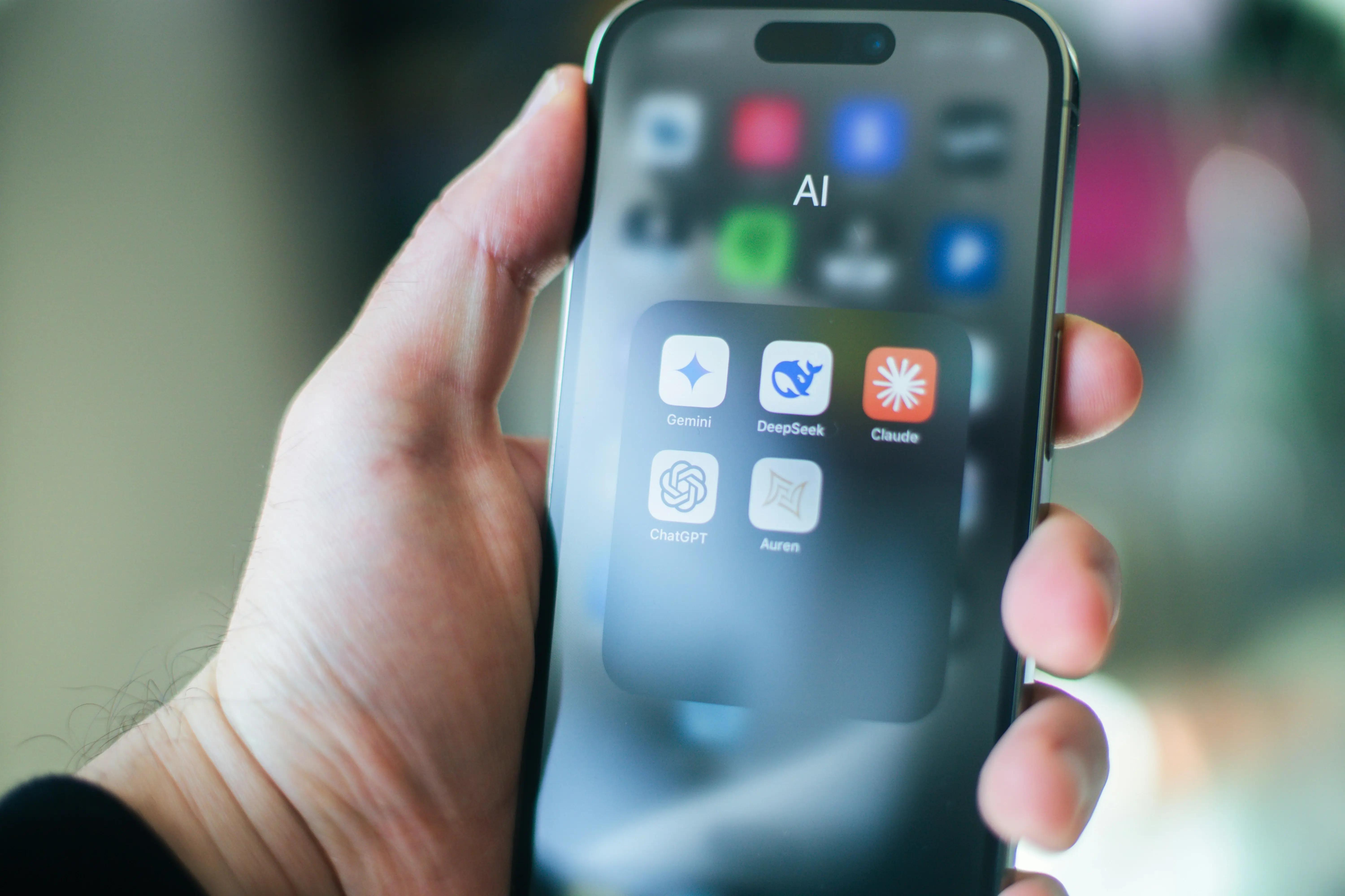 Close-up of a hand holding a smartphone displaying an AI app folder with icons for Gemini, DeepSeek, Claude, ChatGPT, and Auren.