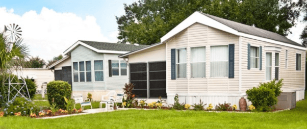 Two mobile homes with beige siding sit side by side on a grassy lawn, showcasing the thoughtful selection of the perfect manufactured home floor plans for your family. Chairs and small plants enhance the front yard, while trees and a windmill grace the background.