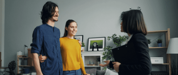In a cozy living room setting, a couple attentively listens to a woman holding a clipboard, sharing top tips for writing Airbnb descriptions that rival Dickens.