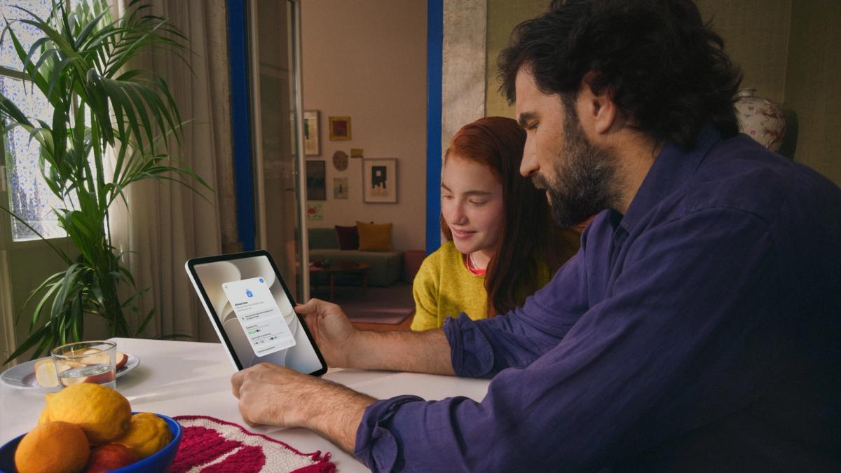 Ein Vater und seine Tochter sitzen gemeinsam am Tisch und richten auf einem iPad die erlaubten Apps für ein Kinderkonto ein