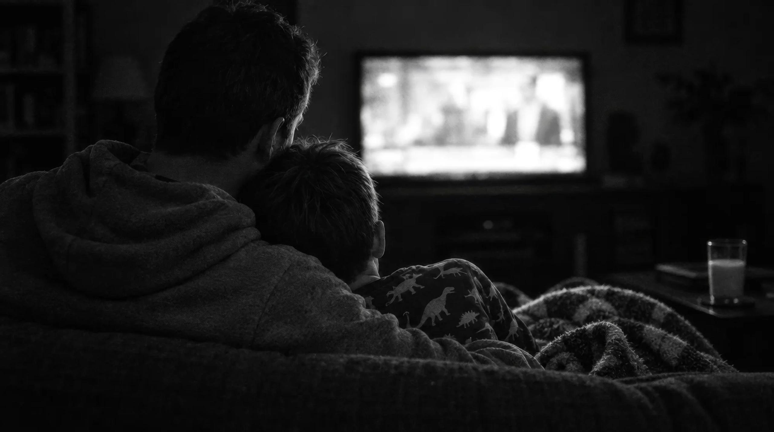 Padre e hijo de espaldas, abrazados en el sofá frente al televisor, en blanco y negro