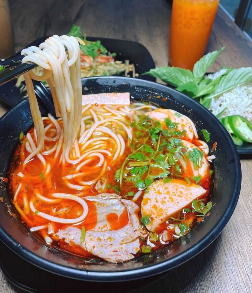 Spicy bún bò Huế noodle soup lifted with chopsticks, with herbs, jalapeños and Thai iced tea