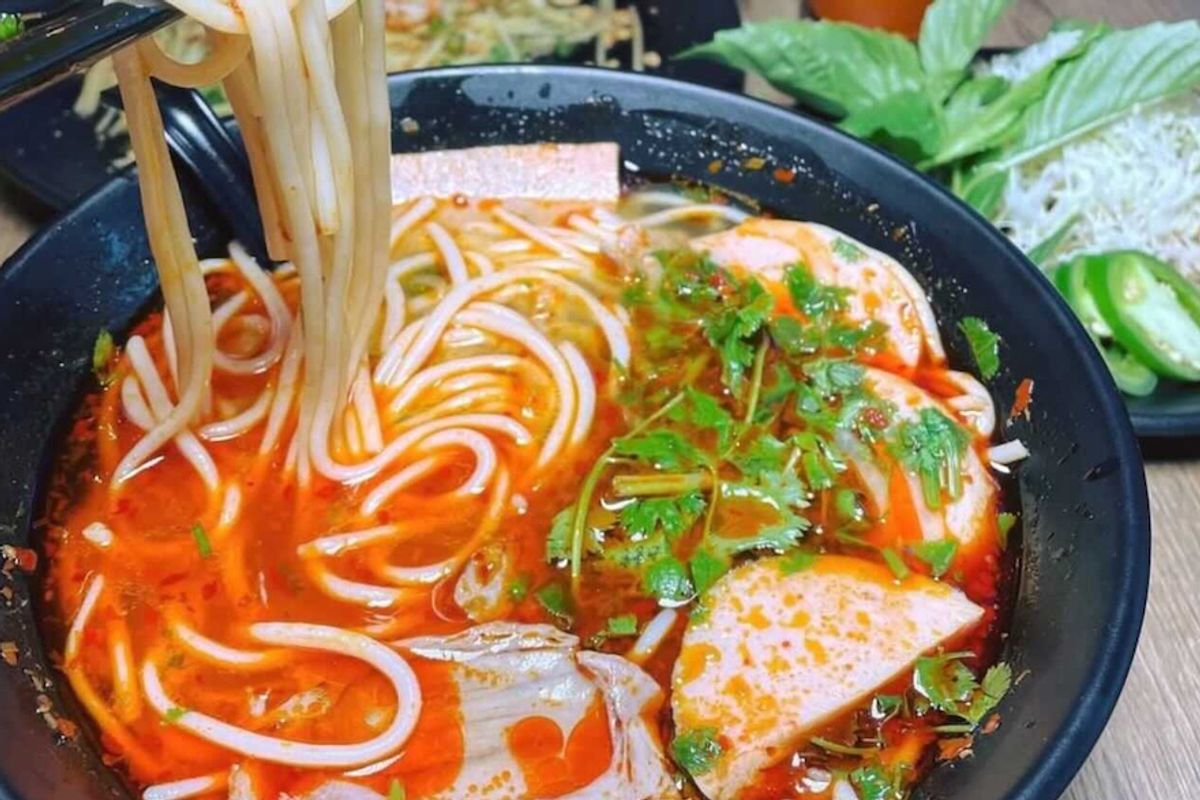 Spicy bún bò Huế noodle soup lifted with chopsticks, with herbs, jalapeños and Thai iced tea