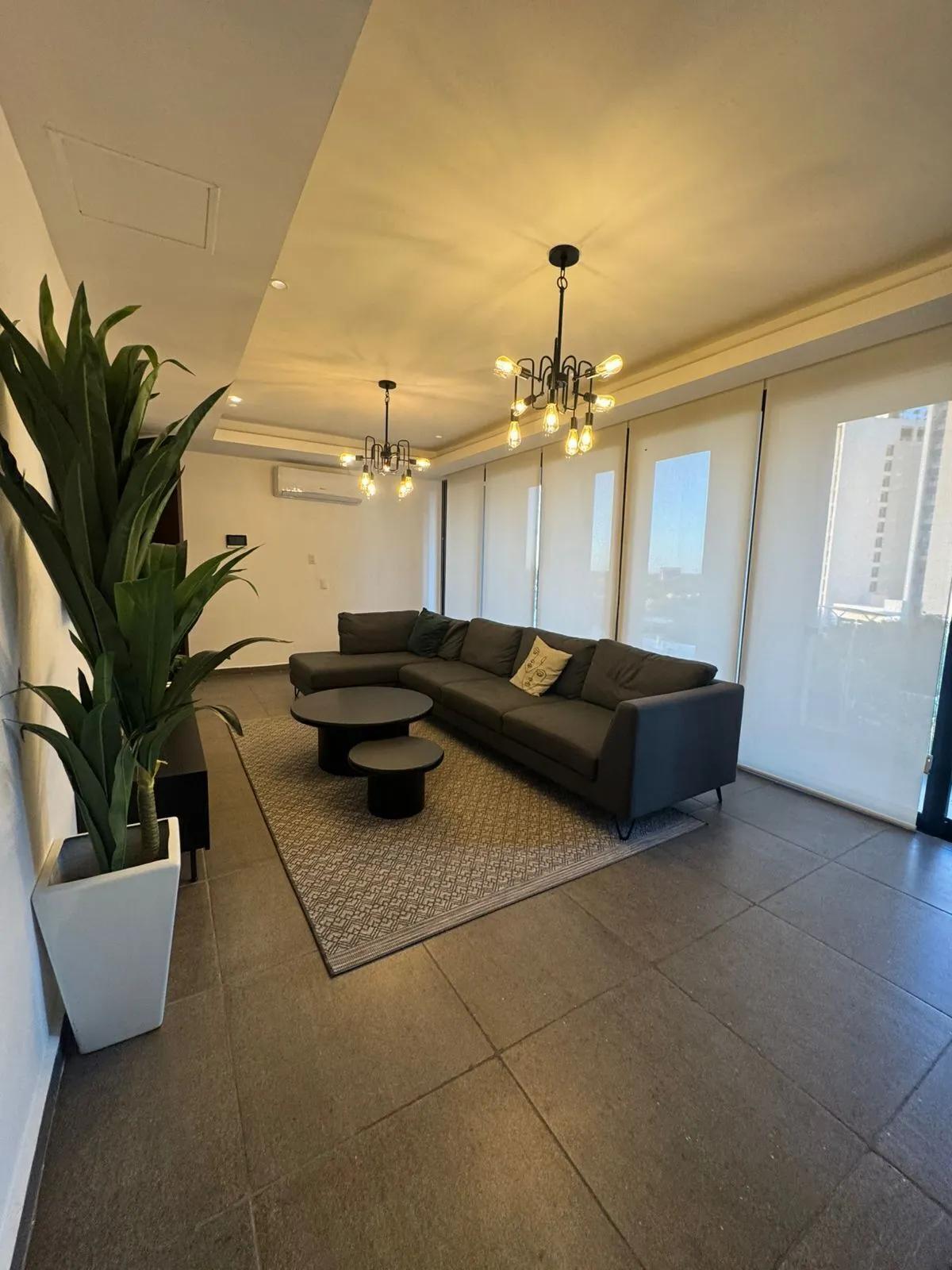 Moderna sala de estar tipo apartamento completamente amueblada. Incluye un sofá seccional gris oscuro, mesas de centro redondas nido y candelabros de estilo industrial. 