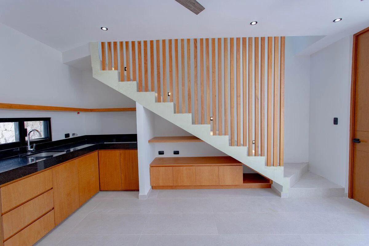 Diseño interior moderno con escalera y cocina. La cocina cuenta con encimeras de granito oscuro y gabinetes de madera clara, que se extienden hasta un banco de almacenamiento incorporado debajo de las escaleras.