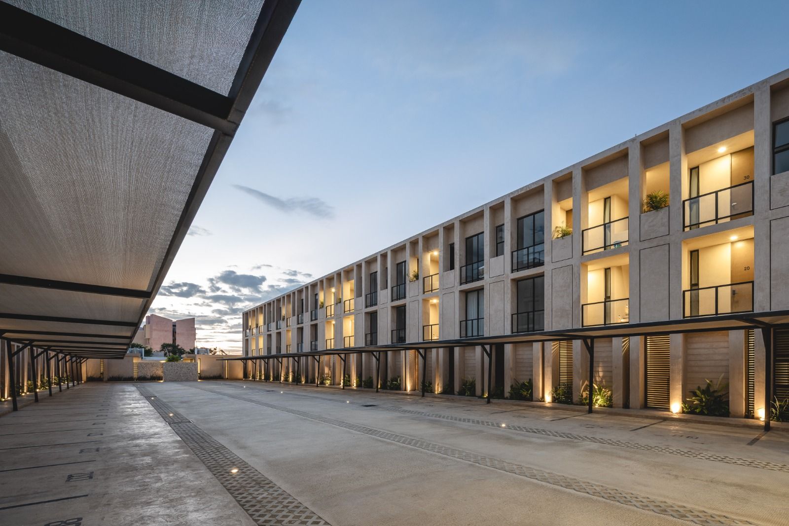 Exterior del complejo habitacional. El diseño presenta una estructura moderna de varios pisos con una fachada de bloques de hormigón y balcones individuales. La planta baja incluye plazas de aparcamiento cubiertas con iluminación contemporánea.