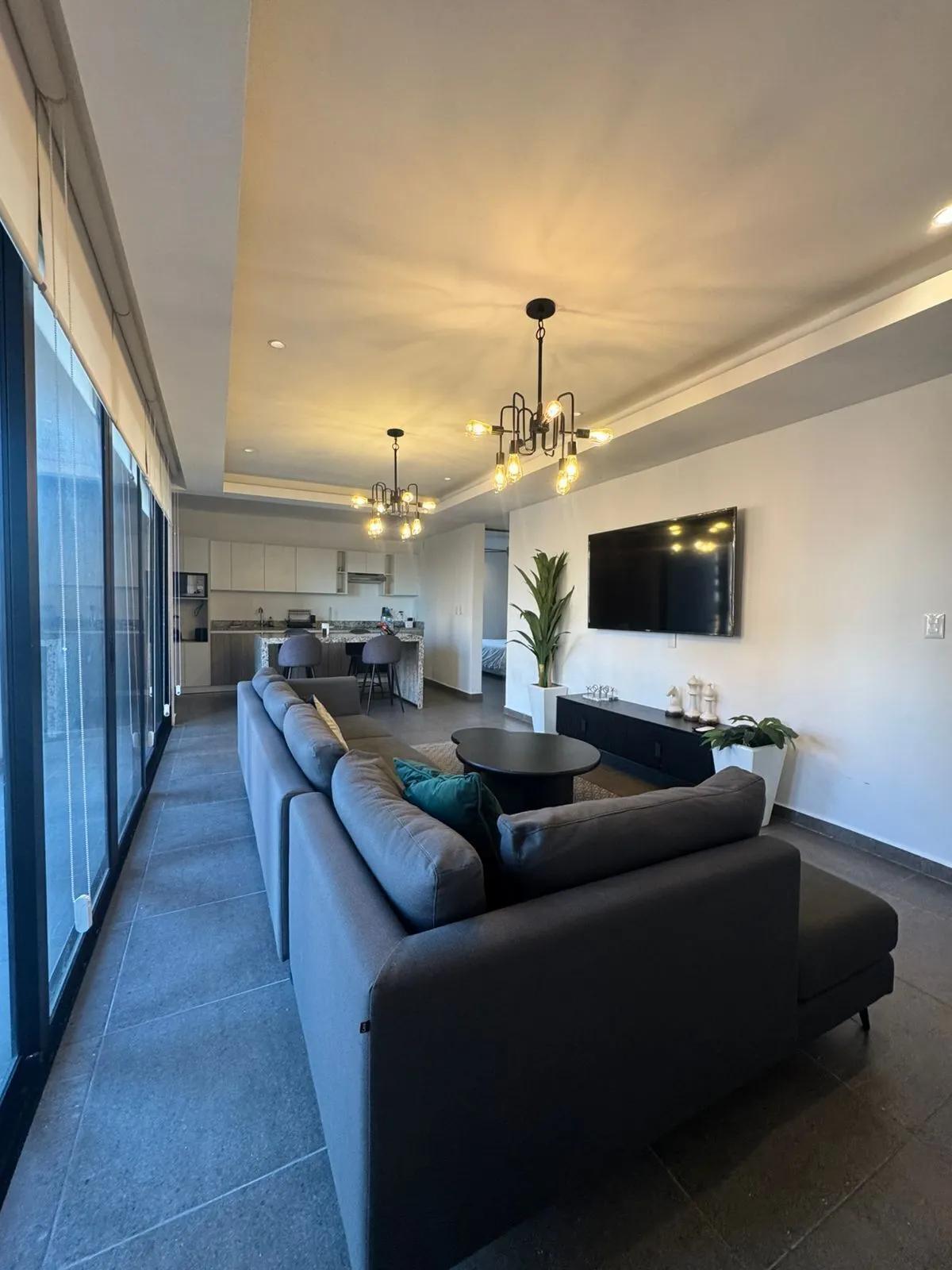 Interior de un departamento con un diseño moderno y abierto que conecta la sala de estar y la cocina. Incluye grandes puertas corredizas de vidrio que conducen a un balcón o terraza, un sofá seccional gris y accesorios de iluminación contemporáneos.