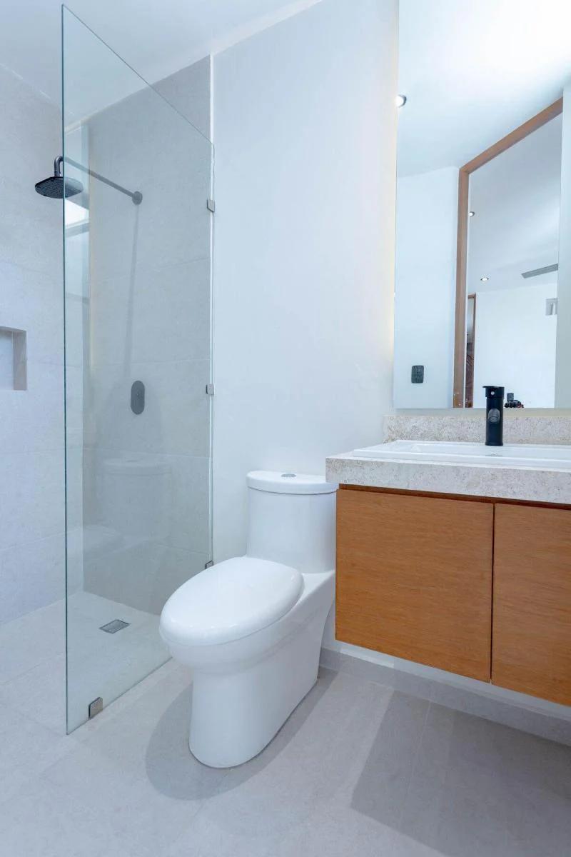 Baño moderno de estética minimalista con paredes blancas y baldosas de colores claros. Hay una cabina de ducha de cristal con un cabezal de ducha de color negro. El tocador incluye gabinetes enchapados en madera, una encimera blanca y un grifo negro contemporáneo.