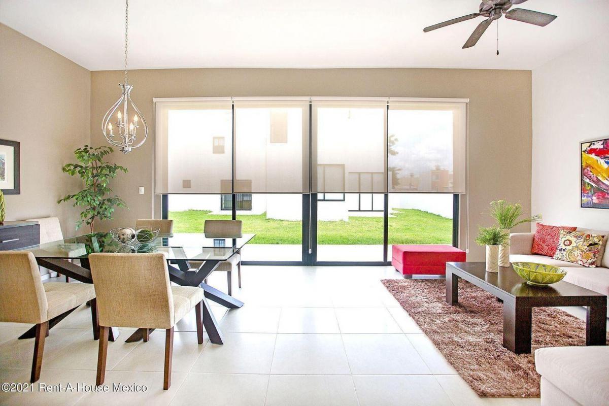 sala de estar y comedor de planta abierta con una decoración moderna, pisos de baldosas y grandes puertas de vidrio que conducen a un espacio exterior con césped. Los muebles clave incluyen una mesa de comedor con tapa de cristal con seis sillas tapizadas, un sofá, una otomana roja y una mesa de café de madera oscura.