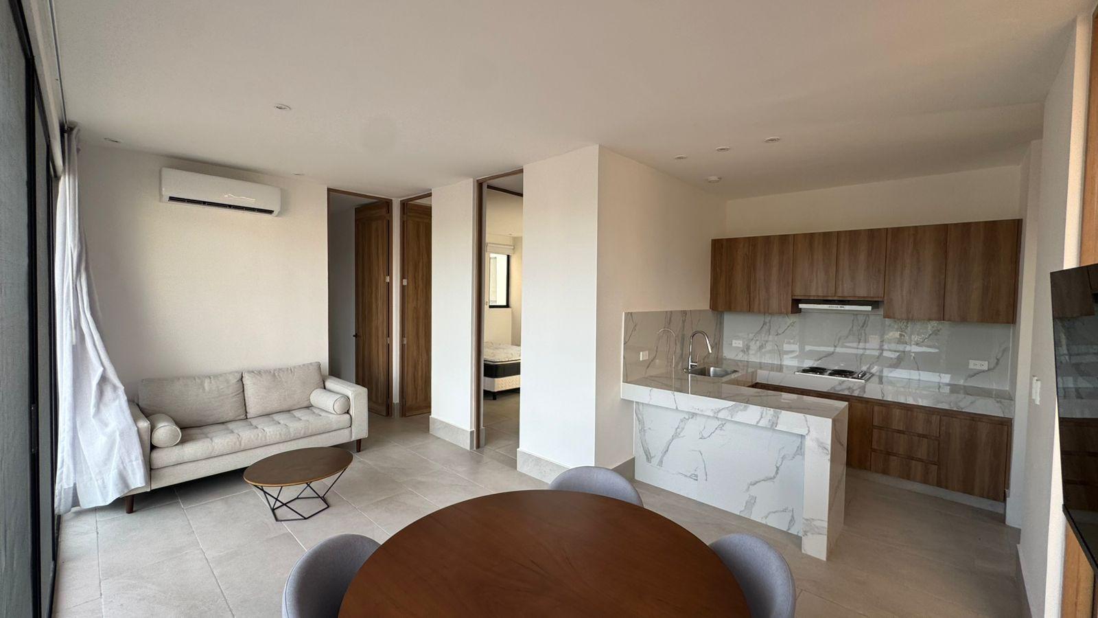 Sala de estar de planta abierta con sofá, mesa de madera, comedor y sillas, y una cocina moderna.